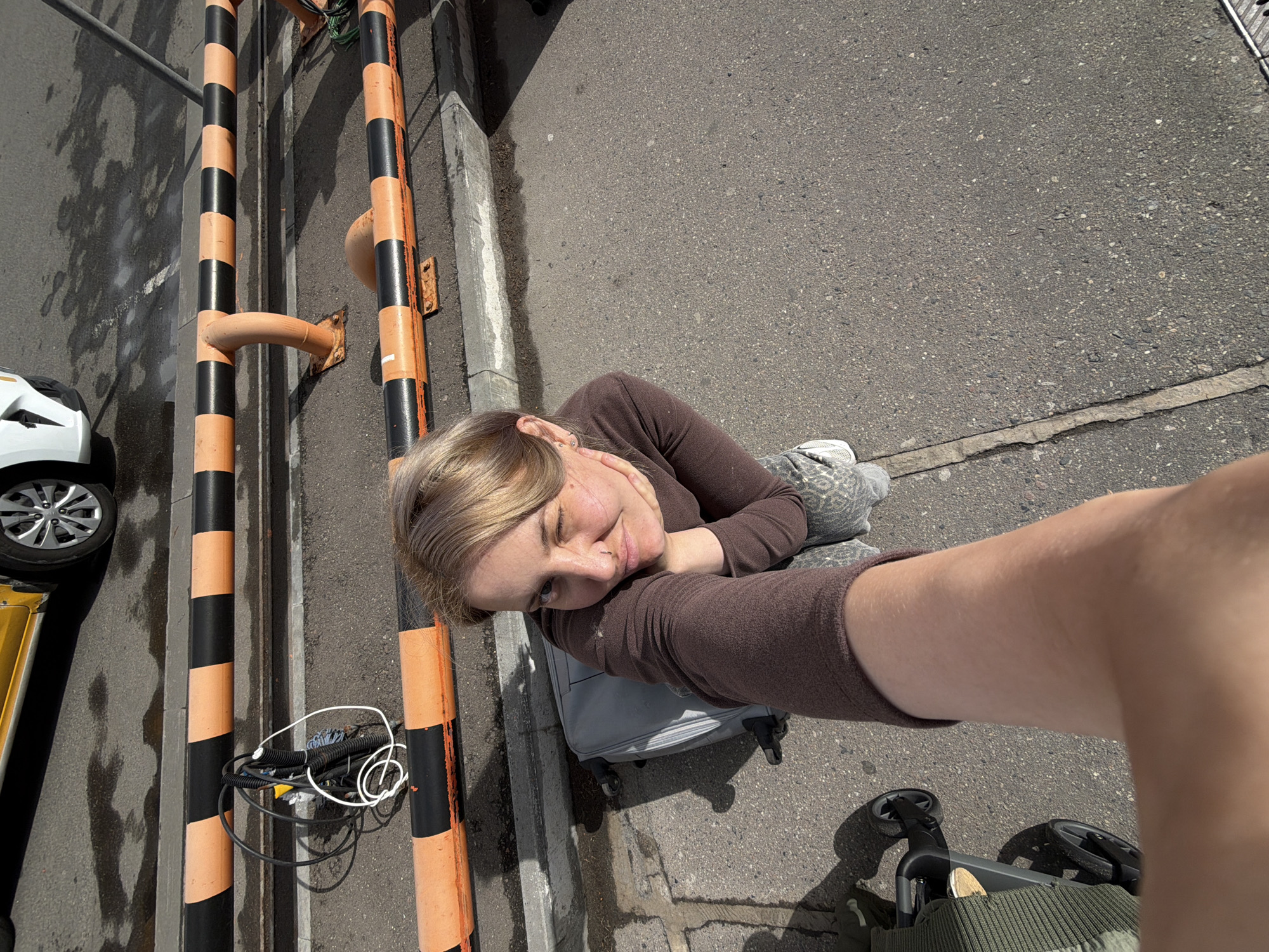 A selfie taken in Domodedovo, Russia, shows a person reclining on a gray suitcase on a concrete surface. The person has fair skin and blonde hair, wearing a brown long-sleeved shirt. They are holding the camera at arm's length. Next to the suitcase are orange and black striped poles, with some cables lying at their base. Adjacent to the poles are a curb, a gutter, and then wet asphalt. To the left is a white car. In the lower right corner, a stroller is visible. The concrete surface is a gray asphalt road.