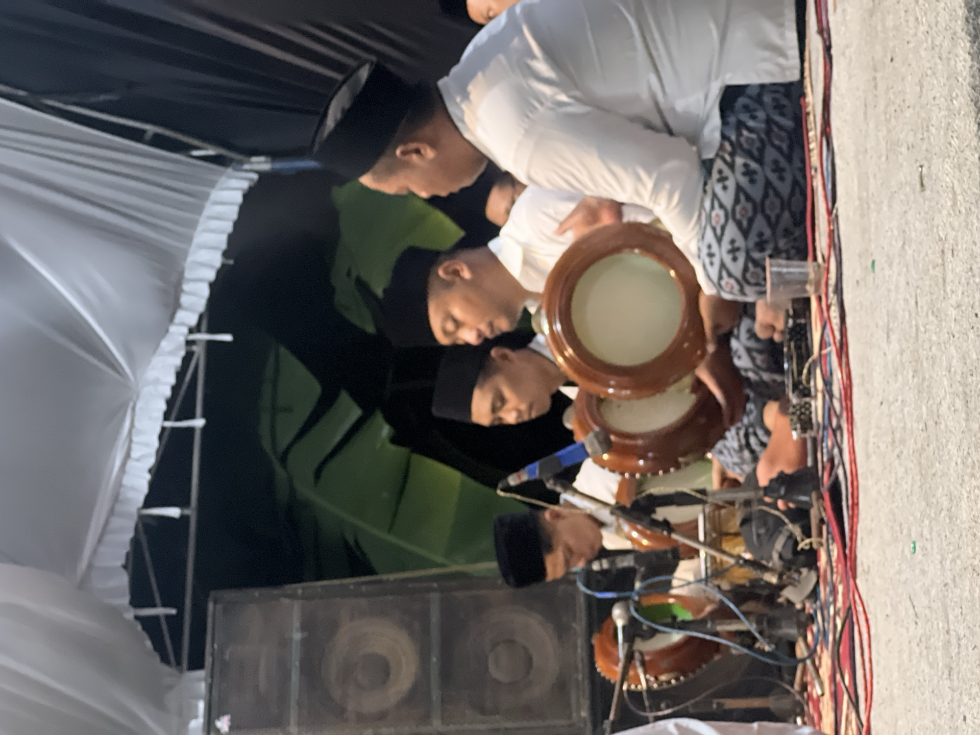 In Wedarijaksa, Indonesia, a group of men wearing black peci hats and white shirts are playing traditional drums. The men are seated on the ground, some are holding the drums, while others are actively playing them. Microphones are positioned in front of the drums to amplify the sound. In the background, large speakers and a white tent with decorative frills are visible. The lighting suggests the scene is taking place at night or in a dimly lit indoor setting. The ground is covered with rugs and cables, indicating a performance or event.