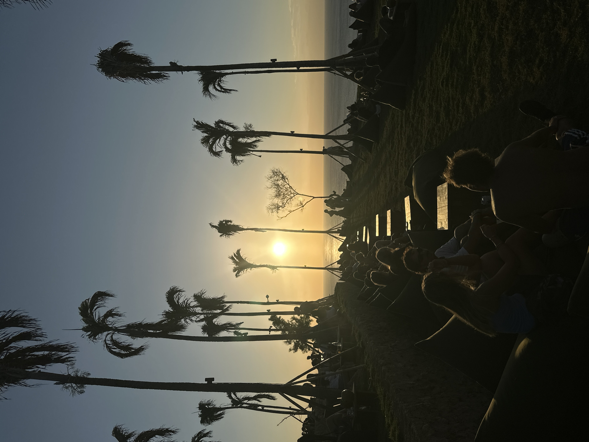 The image captures a sunset scene in Kuta Selatan, Indonesia. A group of people are seated on beanbag chairs and benches on a grassy slope overlooking the ocean. Several palm trees, silhouetted against the bright orange sun, line the edge of the slope. The sun is low on the horizon, casting a warm glow over the water and the sky. The people are casually dressed, and some appear to be in conversation. The overall atmosphere is relaxed and tranquil.