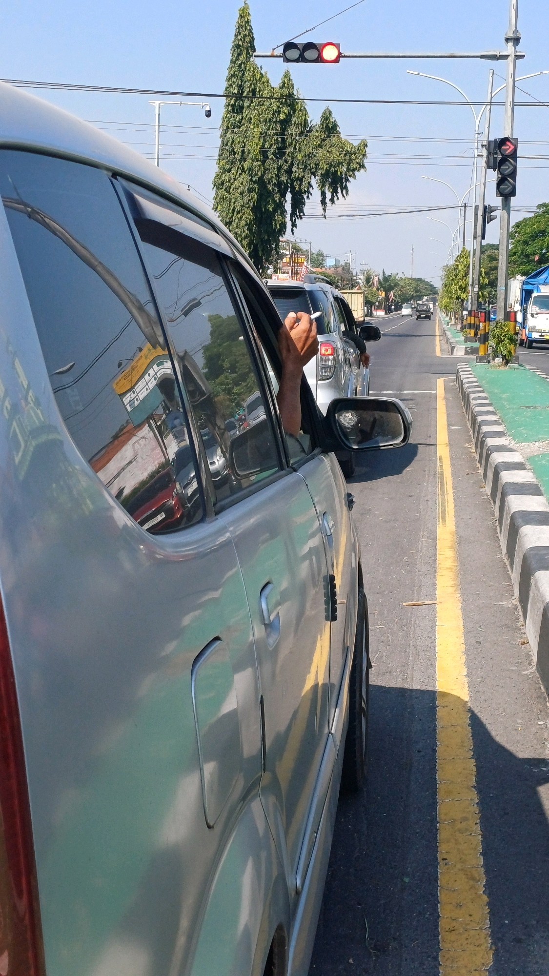 The image depicts a street scene in Jombor, Indonesia, under a clear blue sky. A silver car is in the foreground, viewed from the side, with its rear passenger door open and a person's arm and hand resting on the doorframe, holding a cigarette. The car appears to be stopped in traffic, as suggested by other vehicles visible ahead and a traffic light showing red.
In the background, several cars are lined up on the road. The road is divided by a yellow line and bordered by a curb with a green painted section. Traffic lights are visible on poles, with one showing a red light. Trees with full foliage are present along the roadside. Various buildings and signs are reflected in the window of the silver car. The overall impression is a typical traffic situation on a sunny day in an urban Indonesian environment.