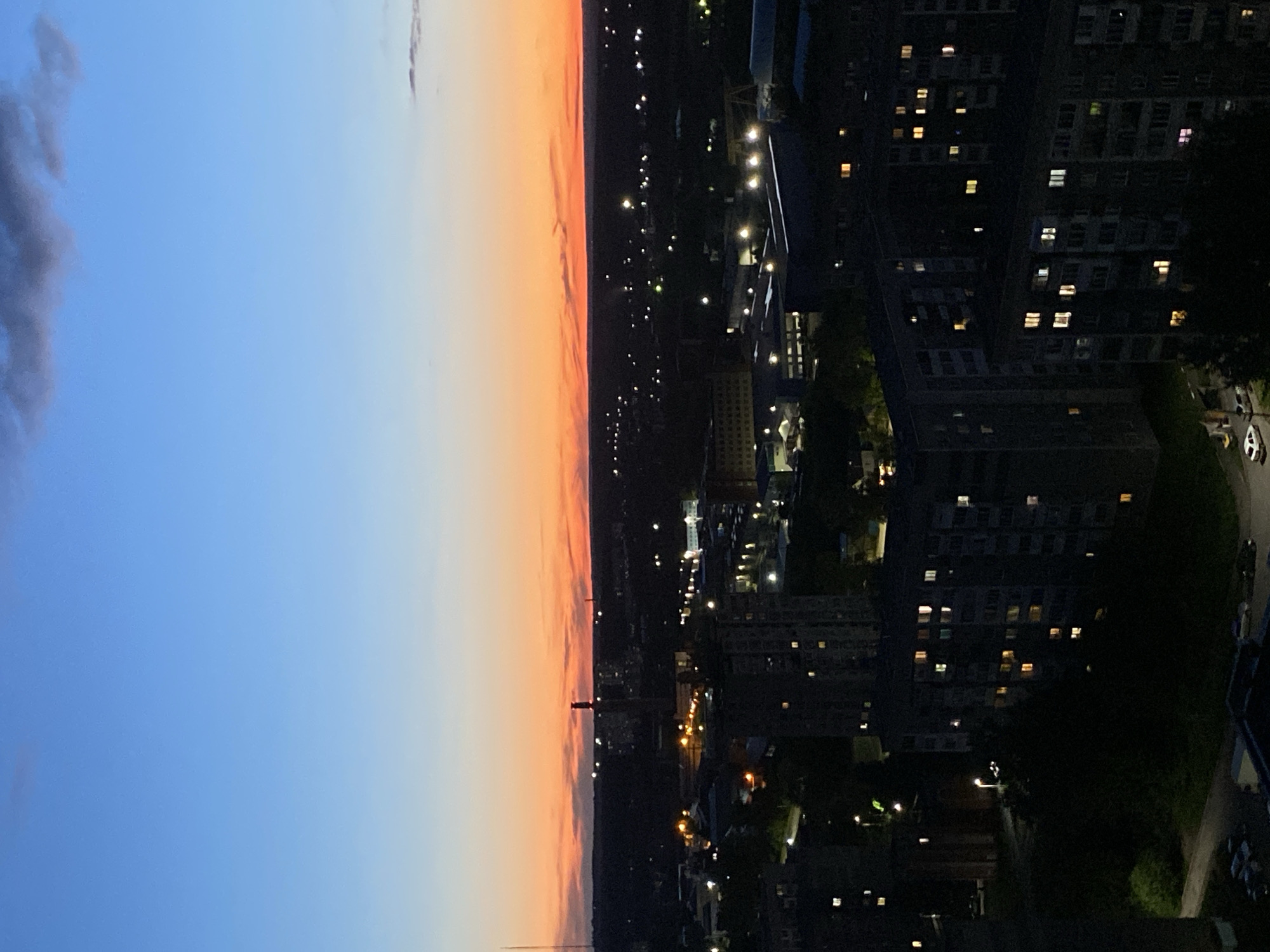 The image presents an expansive urban cityscape at twilight, likely dusk, viewed from a high vantage point. The sky occupies roughly half of the frame, displaying a striking gradient of colors. It transitions from a deep, clear blue at the top left, through lighter blues, to warm oranges and soft pinks along the horizon. A few dark, scattered clouds are visible in the blue portion of the sky.
Below the horizon, the city extends into the distance, with buildings largely silhouetted against the fading light. Numerous multi-story buildings, appearing to be apartment blocks and commercial structures, are scattered across the landscape. Many windows within these buildings are illuminated with varying hues of yellow, orange, and white light, indicating human presence and activity as evening sets in. Pinpricks of light from streetlamps and other urban illuminations dot the dark city, outlining streets and structures.
No specific individuals or overt activities are visible, but the illuminated windows and widespread city lights imply ongoing life within the urban fabric. No discernible text is present in the image. The architectural style, particularly the dense arrangement of residential buildings, aligns with typical urban environments found in Izhevsk, Russia, as per the provided location context. The scene evokes a sense of the city transitioning into night, peaceful yet vibrant with internal activity.