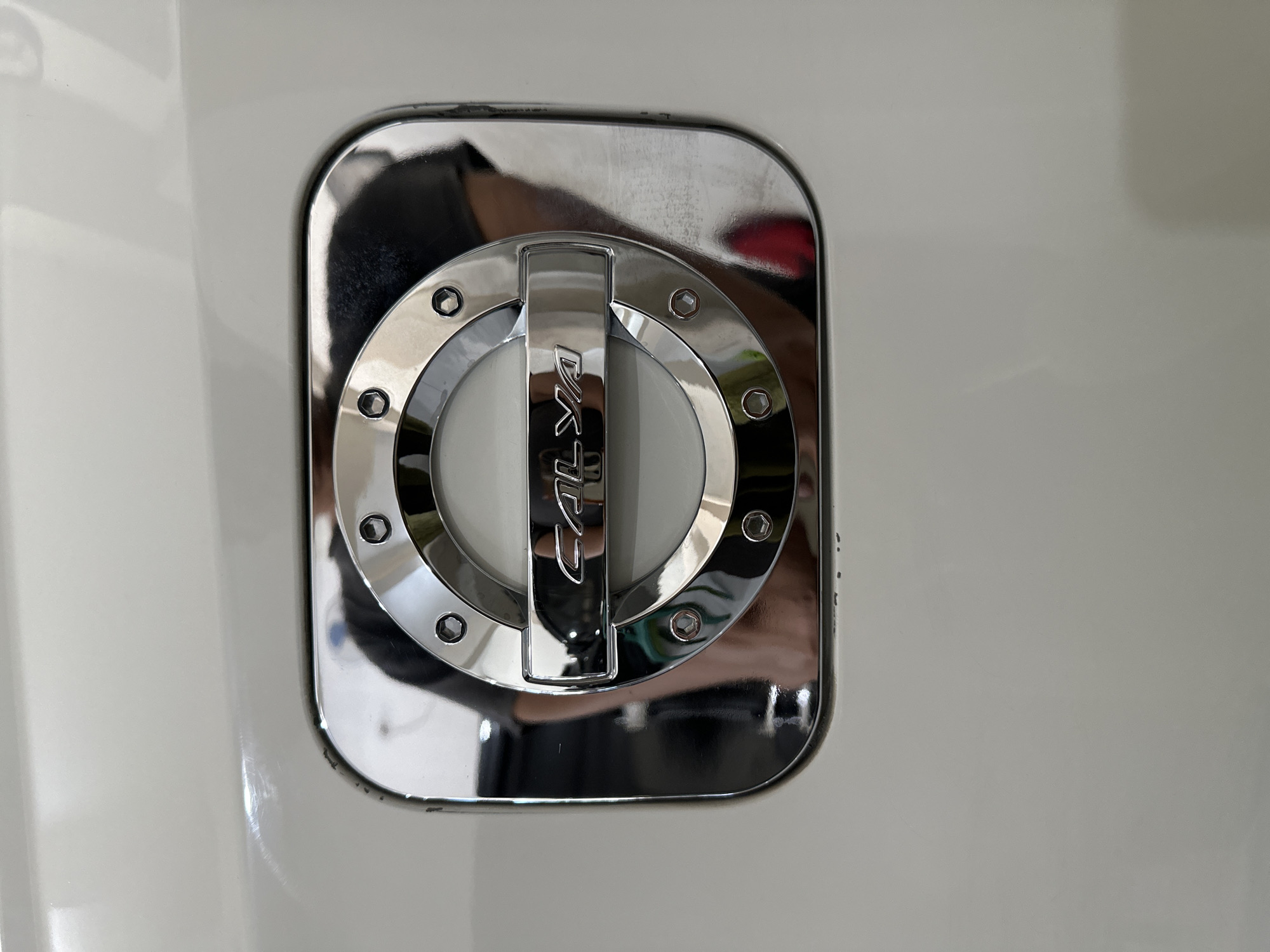 The image shows a close-up of a chrome fuel filler cap on a white vehicle. The cap is circular with a decorative outer ring and a central element featuring the word "CALYA" in raised chrome lettering. The fuel cap is slightly recessed into the vehicle's bodywork, which appears to be a slightly off-white or cream color. There are reflections on the chrome surface of the cap, indicating the presence of other objects or people nearby, possibly including a person wearing a dark t-shirt. The surrounding vehicle body shows some light scuffing and minor imperfections. The image is taken from a slightly elevated, front-on perspective. There are no indications or context linking this image to Bulakrejo, Indonesia.
