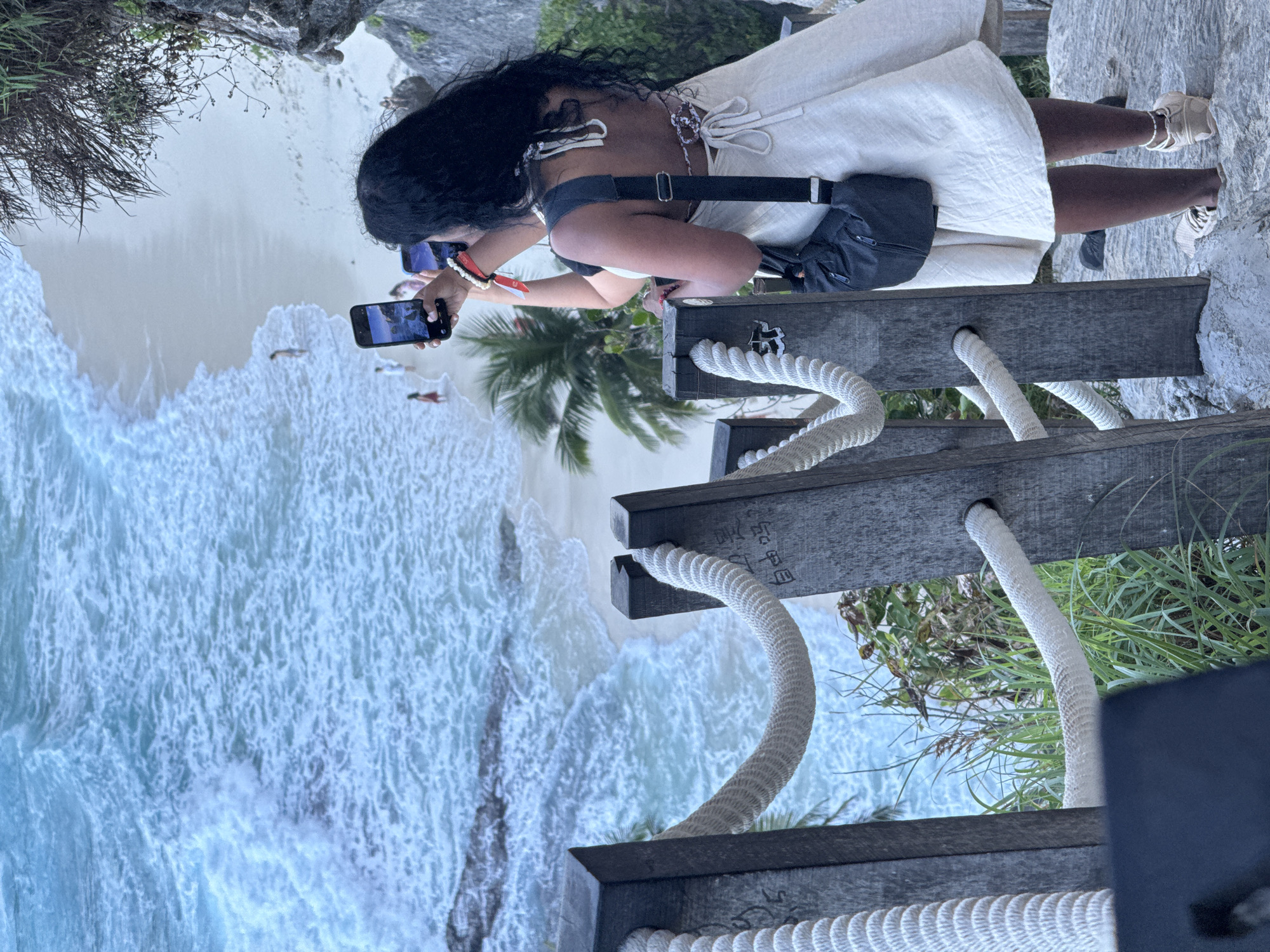 A woman with long, dark, curly hair stands on a rocky path, facing away from the camera, taking photos of a beach with her cell phone. She is wearing a white, halter-neck dress and a dark crossbody bag. A white rope railing guides the path. The ocean waves crash onto a sandy beach where a few people can be seen. This scene is likely at Diamond Beach in Nusa Penida, Indonesia, a popular tourist destination known for its stunning cliffs and turquoise waters.