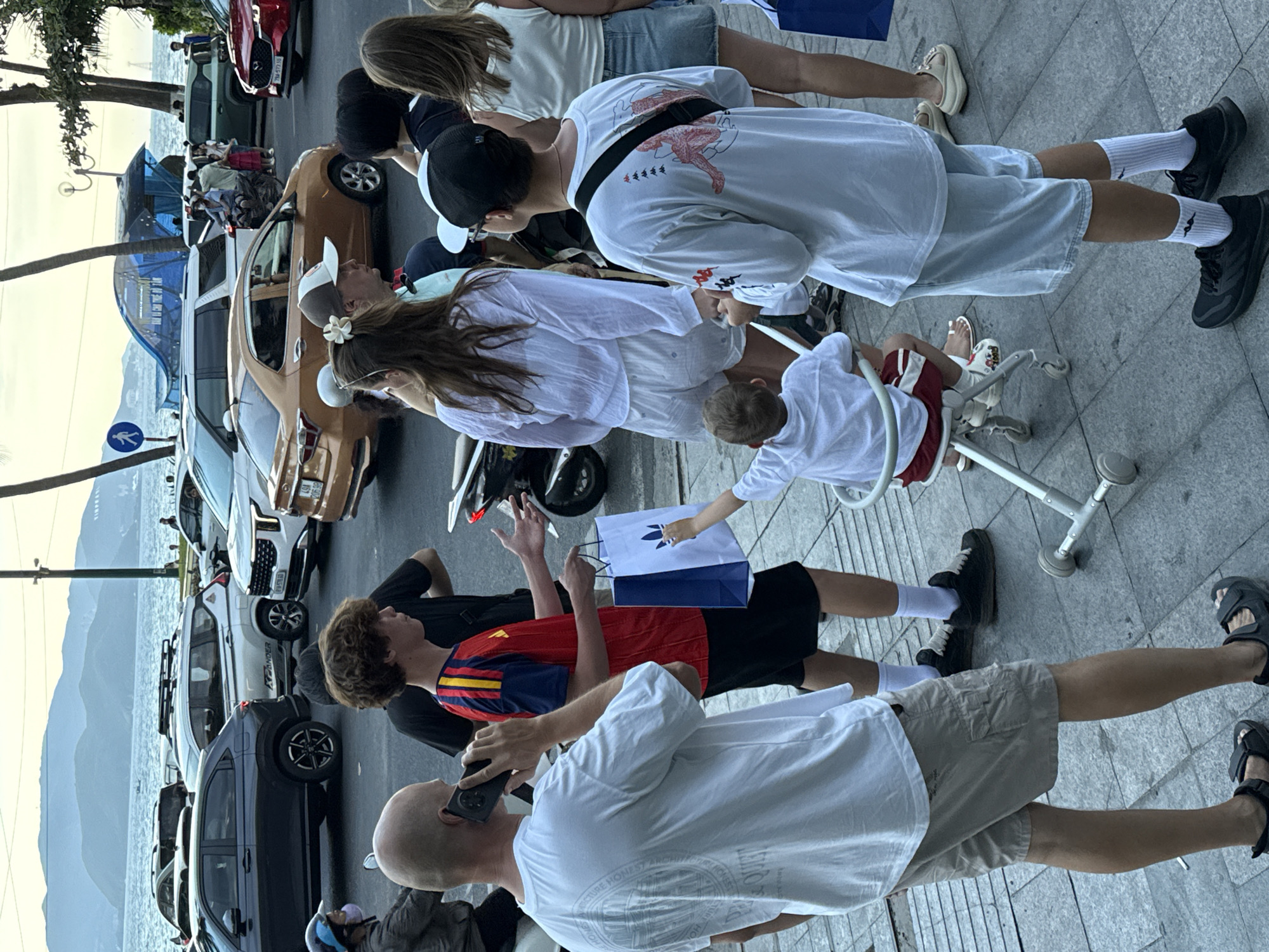 A diverse group of people are gathered on a sidewalk near a street in Nha Trang Ward, Vietnam. The scene appears to be a busy urban environment, possibly near a coastal area given the presence of water and mountains in the background. A man in the foreground is on the phone, wearing a white t-shirt and khaki shorts. Beside him, a young person in a red and yellow shirt holds a blue shopping bag with an Adidas logo. In the mid-ground, a woman with a flower in her hair, wearing a white top and shorts, is with another person. Further back, a child is seated in a stroller-like device. Various vehicles, including cars and a motorcycle, are visible on the street, suggesting traffic. Palm trees line the sidewalk, and a blue structure with a whale graphic is visible in the distance. The lighting suggests it is either early morning or late afternoon.