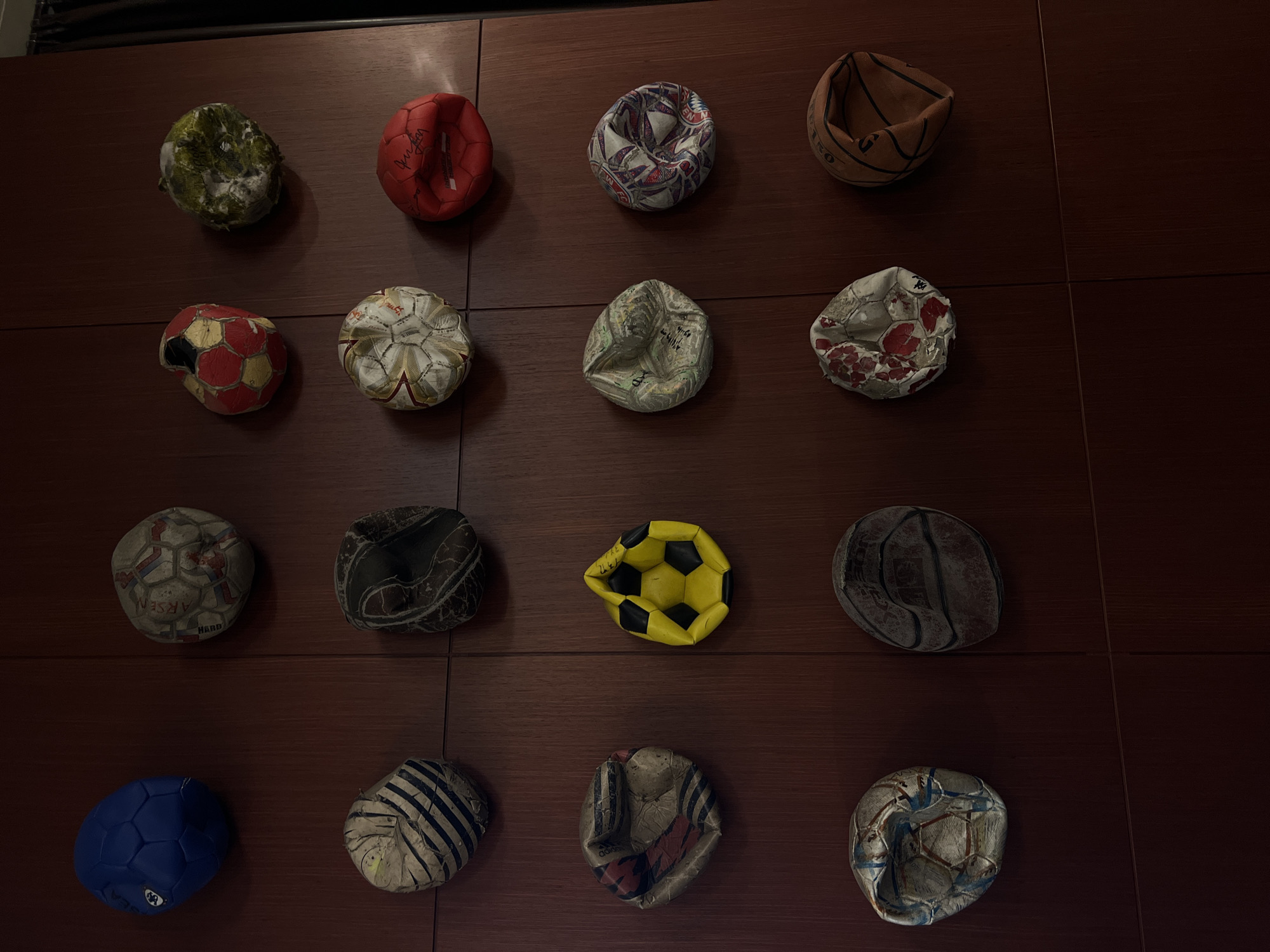 The image depicts a collection of deflated or worn-out sports balls arranged on a dark brown surface with a grid pattern, possibly a wall or paneling. There are 16 balls in total, organized in a 4x4 grid. The balls appear to be various types, including soccer balls, basketballs, and possibly other kinds of sports balls. The objects are arranged at seemingly equal distance. All balls show signs of significant wear, damage, and deflation. The balls vary in color and design, with some featuring recognizable patterns like the black and white panels of a soccer ball or the stripes of a basketball. The yellow ball is in the 3rd row of balls and 3rd ball of that row. The environment is indoor, with artificial lighting and a dark brown background. The location is unknown, but could be the city of Yerevan, Armenia. There are no people in the image, and no actions or interactions visible.