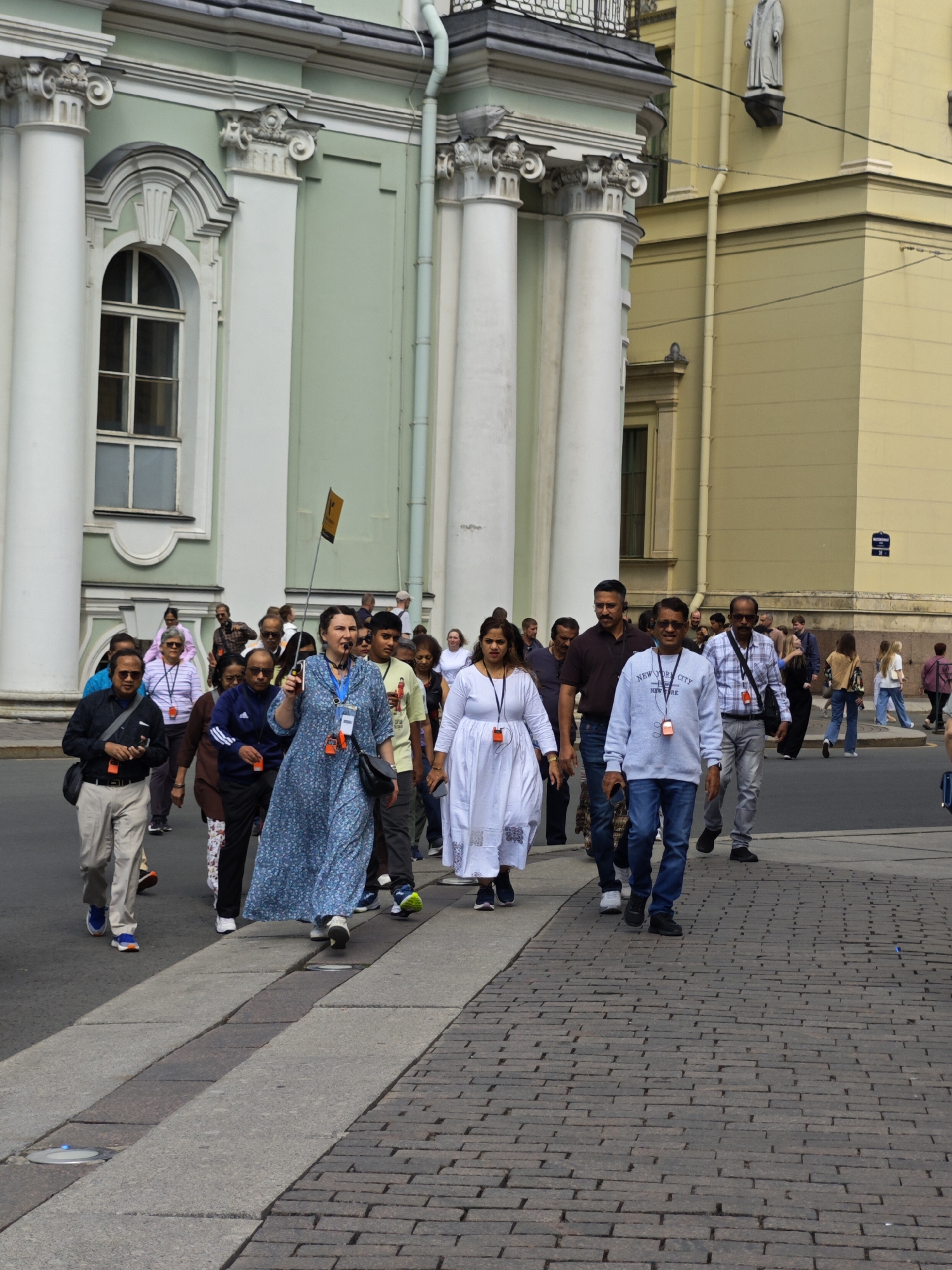 A group of tourists is walking on a cobblestone street in front of a pale green building with white columns and arched windows. A woman in a blue floral dress is leading the group, holding a flag on a stick. She is wearing a lanyard with an ID badge. Several other individuals in the group are also wearing lanyards with badges. The people are dressed in casual clothing, and the weather appears to be overcast. Some of the tourists are carrying bags or wearing backpacks. The building has classical architectural features, including a statue on the upper facade of an adjacent yellow building.