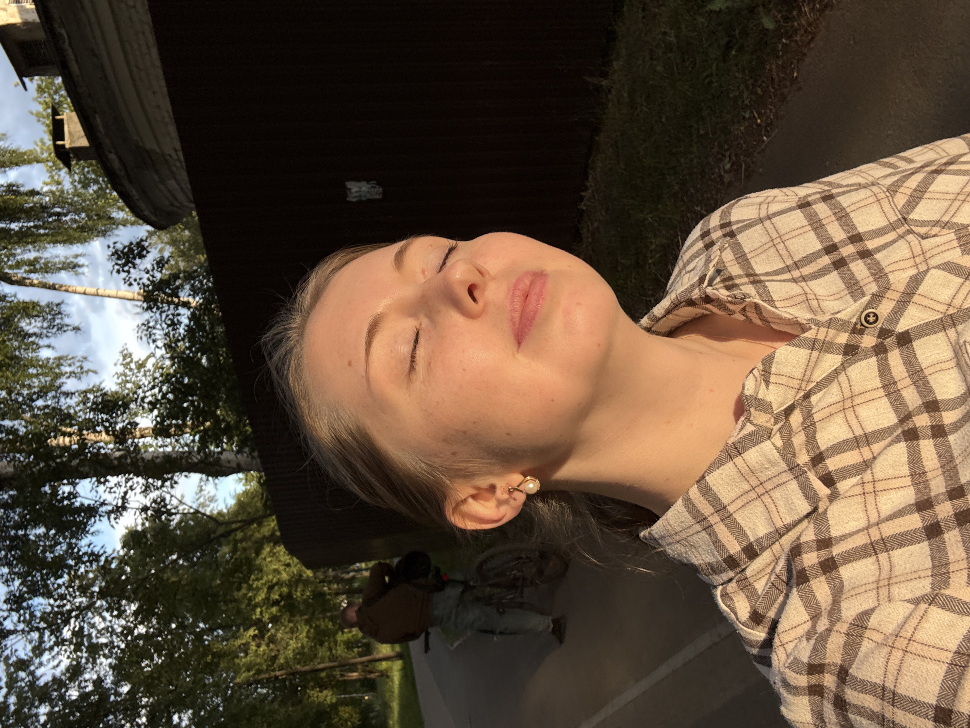 A young woman with closed eyes and a gentle smile is captured in a close-up selfie, bathed in the warm glow of the setting sun. She wears a light-colored plaid shirt. In the background, a person rides a bicycle down a paved path, with a dark wooden fence and trees forming the backdrop. A building with a rounded structure and a chimney is visible above the fence. The scene appears to be set in a park or a suburban area.