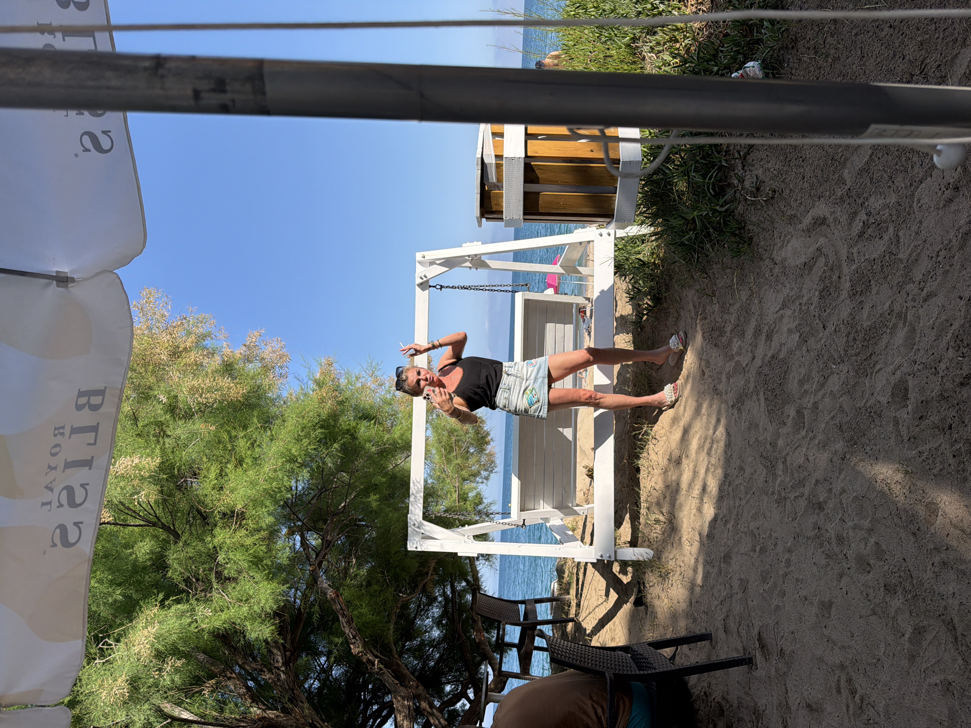 The image depicts a beach scene in Mont-roig del Camp, Spain, during daylight hours. A woman stands next to a white swing set on a sandy beach, holding a cigarette. She is wearing sunglasses, a black tank top, and a denim skirt. The ocean is visible in the background. To the left, a tree provides partial shade, and a beach umbrella is also present. In the foreground, a few chairs are situated on the sand. Behind the swing set is a wooden structure, and vegetation grows alongside the beach area.