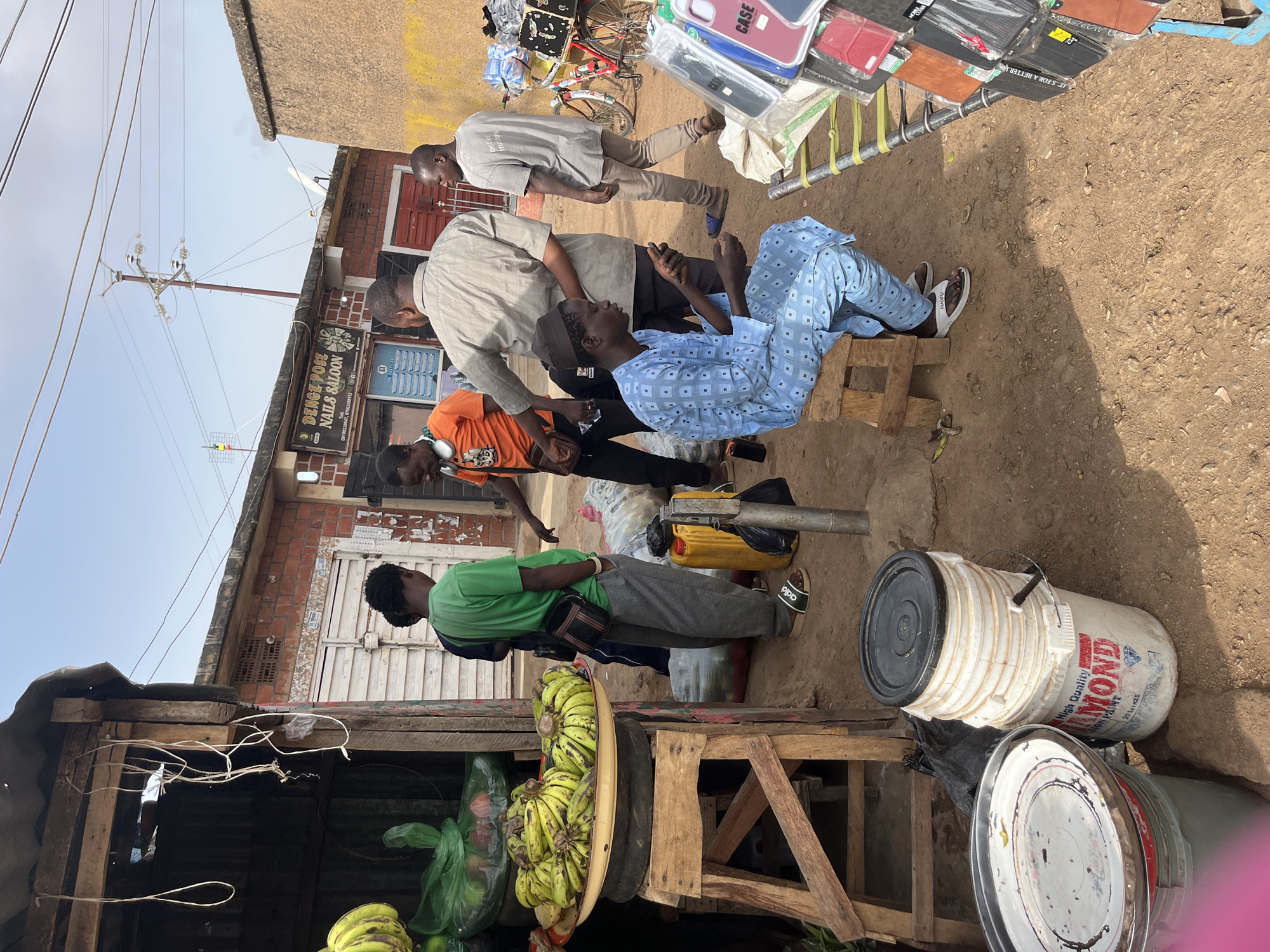 The image shows a street scene in Gura Top, Nigeria. There are several people present, engaged in various activities.
One man is seated on a wooden stool wearing a light blue patterned outfit. Three other men are standing near him, possibly in conversation. One man is wearing a grey shirt and the second one is wearing an orange shirt. A fourth man is standing with a green shirt.
A small storefront is visible, with a sign that reads "DENGE POSE NAILS SALOON."
In the foreground, there is a fruit stand with bananas and other items. Buckets of paint are also visible. The ground is dirt and there are various items scattered around. Above the scene, power lines are stretched across the sky.
To the right, a man is walking with a bicycle and a rack of phone cases is displayed.