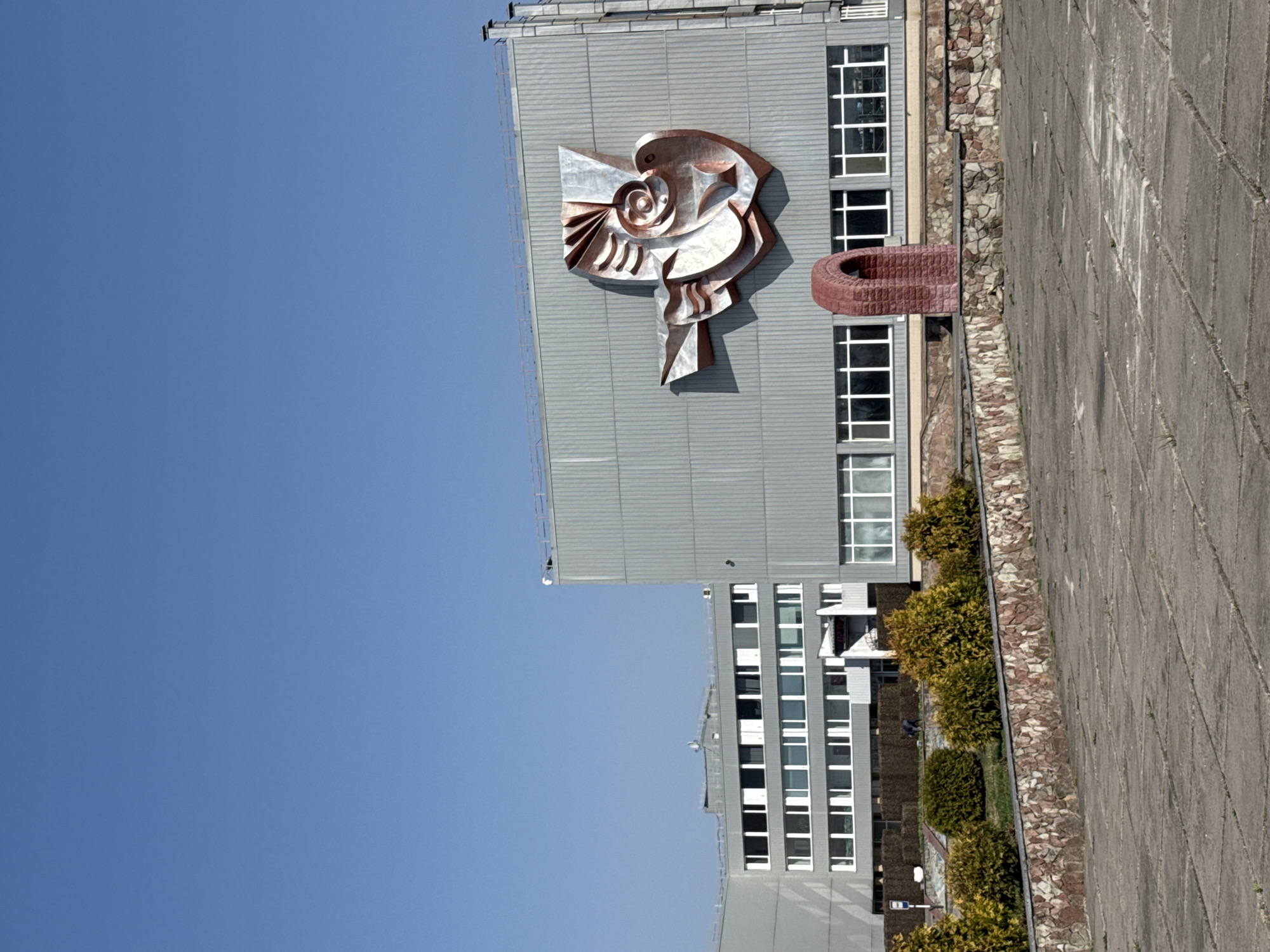 The image shows a gray building in Pryp'yat', Ukraine. On the side of the building is a large, metallic sculpture. There are rows of windows along the side of the building. A concrete path runs alongside the building, separated by a strip of bushes and rocks. The sky is clear blue.