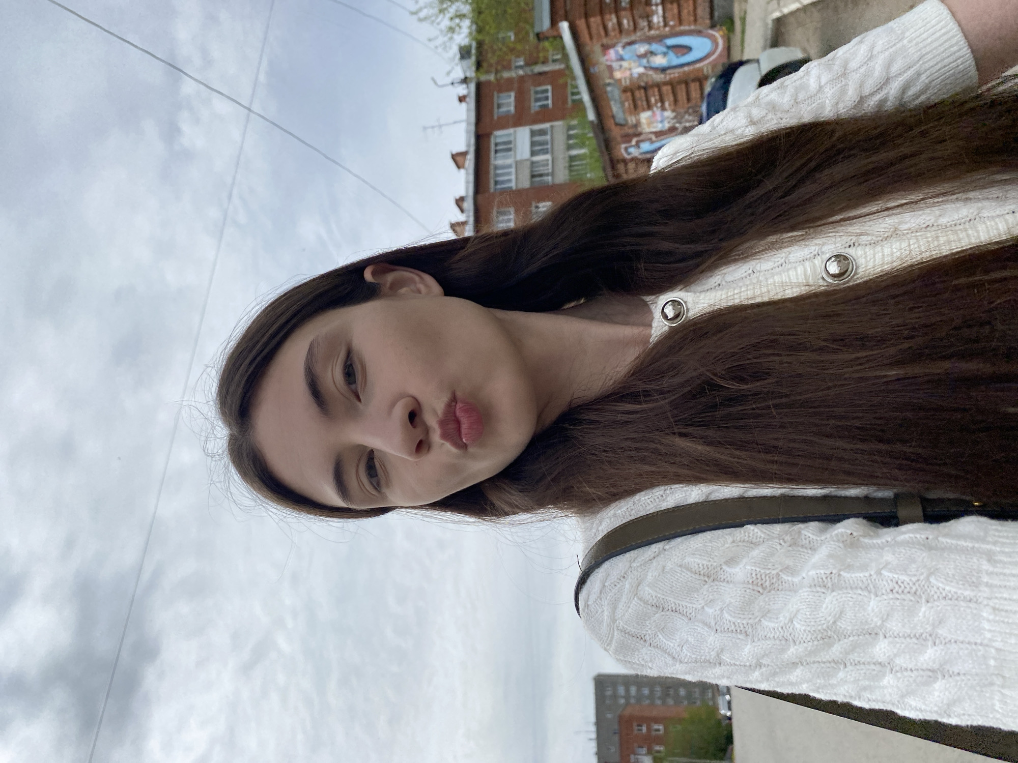 A selfie taken outdoors shows a young woman with long, dark brown hair, wearing a white cable-knit sweater with decorative buttons and a dark-colored shoulder bag. She is making a "duck face" expression, puckering her lips. The background includes a cloudy sky, buildings, and trees. One of the buildings appears to be made of red brick and features a mural. Buildings appear to be in Votkinsk, Russia.