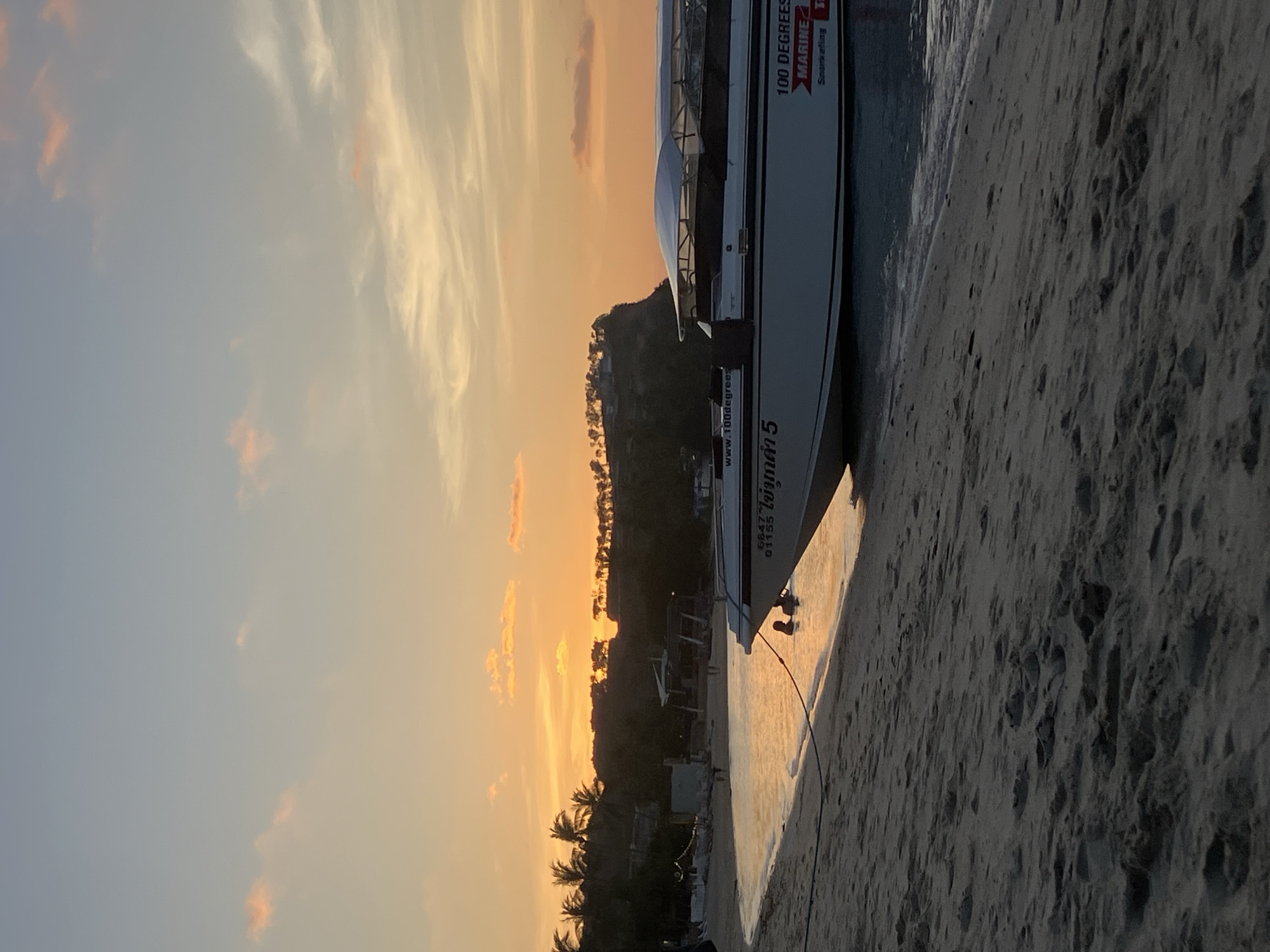 A sunset scene is depicted on a sandy beach in Baan Bang Rak, Thailand. The sky displays a gradient from bright orange and yellow at the horizon to pale blue and grey overhead, with scattered, thin clouds reflecting pink and orange light. In the mid-ground, a dark, tree-covered hillside or cliff is silhouetted against the setting sun, with smaller trees and discernible lights on its summit. Palm trees are visible on the left side of the distant landmass, alongside indistinct beach structures and figures walking on the shore.
In the foreground and mid-ground, a white and dark-colored speedboat or tour boat, marked with "6647 01155", "ไอดอลค้า 5" (Thai script), "www.100degrees.com", and "100 DEGREES MARINE Snorkeling", is partially beached in the shallow, sun-reflective water. Its bow points towards the left, and a dark rope extends from it into the water. Two individuals are partially submerged in the water near the boat's bow, with their heads and shoulders visible. The immediate foreground consists of light-colored sand, showing footprints and small irregularities, with gentle waves lapping at the shore.
