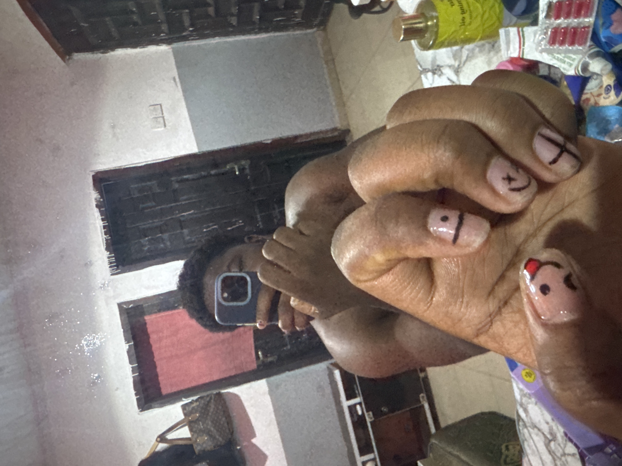 A person is taking a selfie in a mirror. The person has their hand in the foreground, with painted nails with smiley face and cross designs. In the background is a room with a window covered by bars and a curtain. Some bags are located under the window. Various items are cluttered on a table in the background. The location is potentially Gbongan, Nigeria, but there is not sufficient evidence to confirm this.