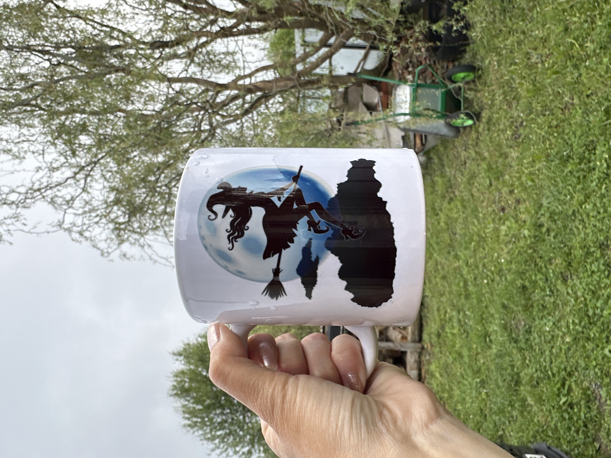 A person holds a white coffee mug with a Halloween-themed design, depicting a witch riding a broom in front of a full moon, with a castle silhouette on the side. The background shows a grassy area with trees and some gardening equipment. The location is likely in Среднее Кечево, Russia.