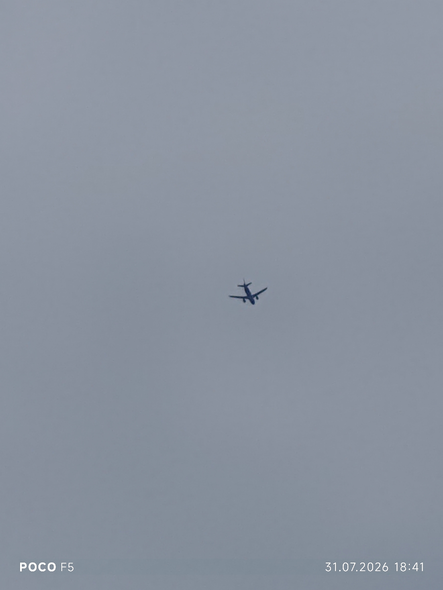 The image shows an airplane in flight against a uniformly overcast sky. The airplane is small and silhouetted against the lighter sky, making it difficult to discern specific details. The sky appears to be a pale gray, indicating cloudy weather. In the bottom left corner of the image, the text "POCO F5" is visible, likely a watermark from the device used to capture the photo. In the bottom right corner, the date "31.07.2026" and time "18:41" are displayed. There are no other discernible objects or subjects in the frame, and no information within the image suggests a connection to Camogli, Italy.