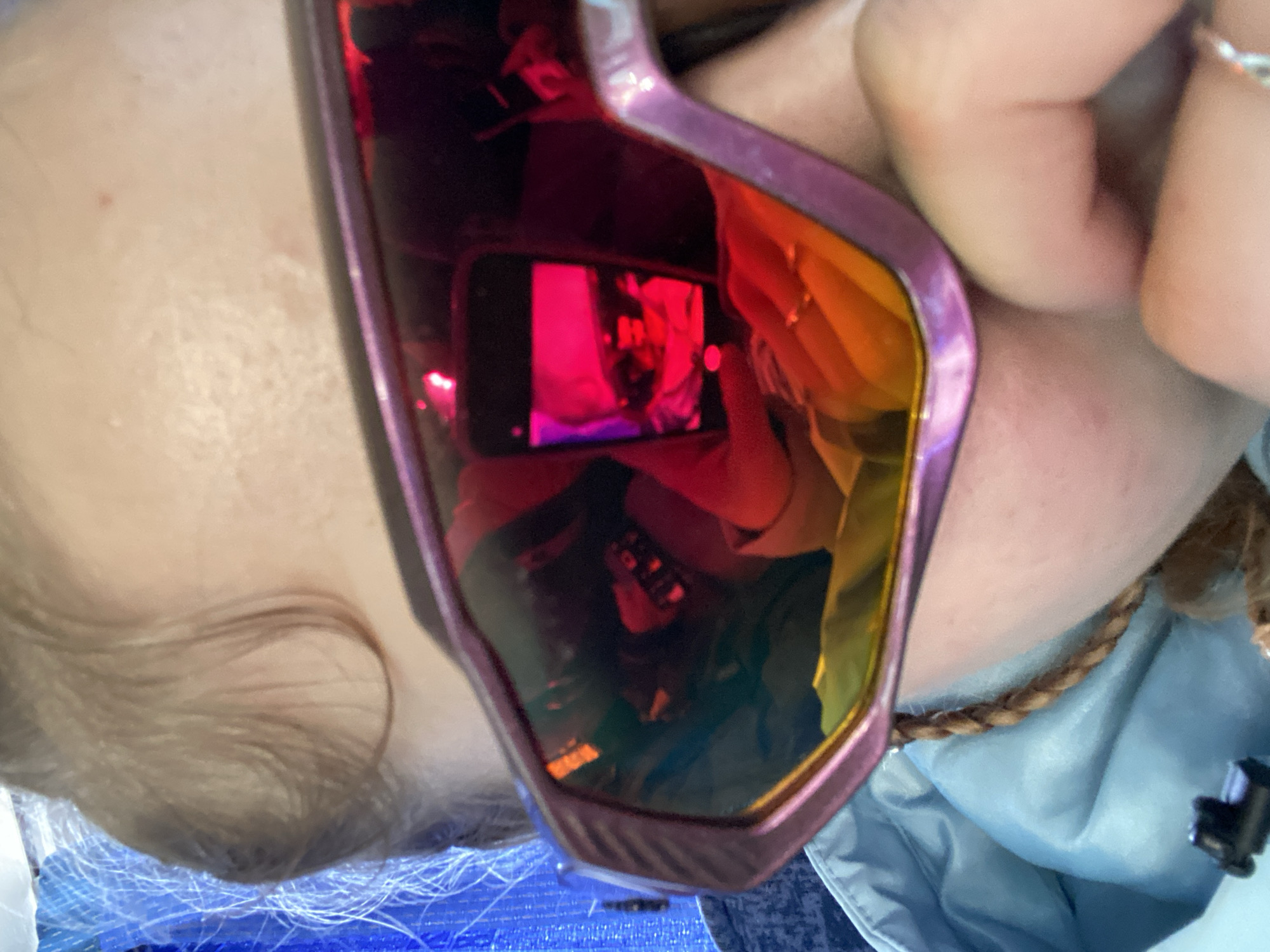 The image is a selfie taken in reflective ski goggles. The goggles have a pink frame and the reflective lenses show a reflection of a person holding a mobile phone, presumably taking the picture. The phone screen in the reflection shows a red-toned image. The person wearing the goggles has light skin, blonde hair, and is wearing a light blue jacket or top with a brown cord detail. The background visible beneath the goggles suggests a blue netting or fabric. There is no information in the image or provided that specifies a location within the city of Berezniki, Russia.