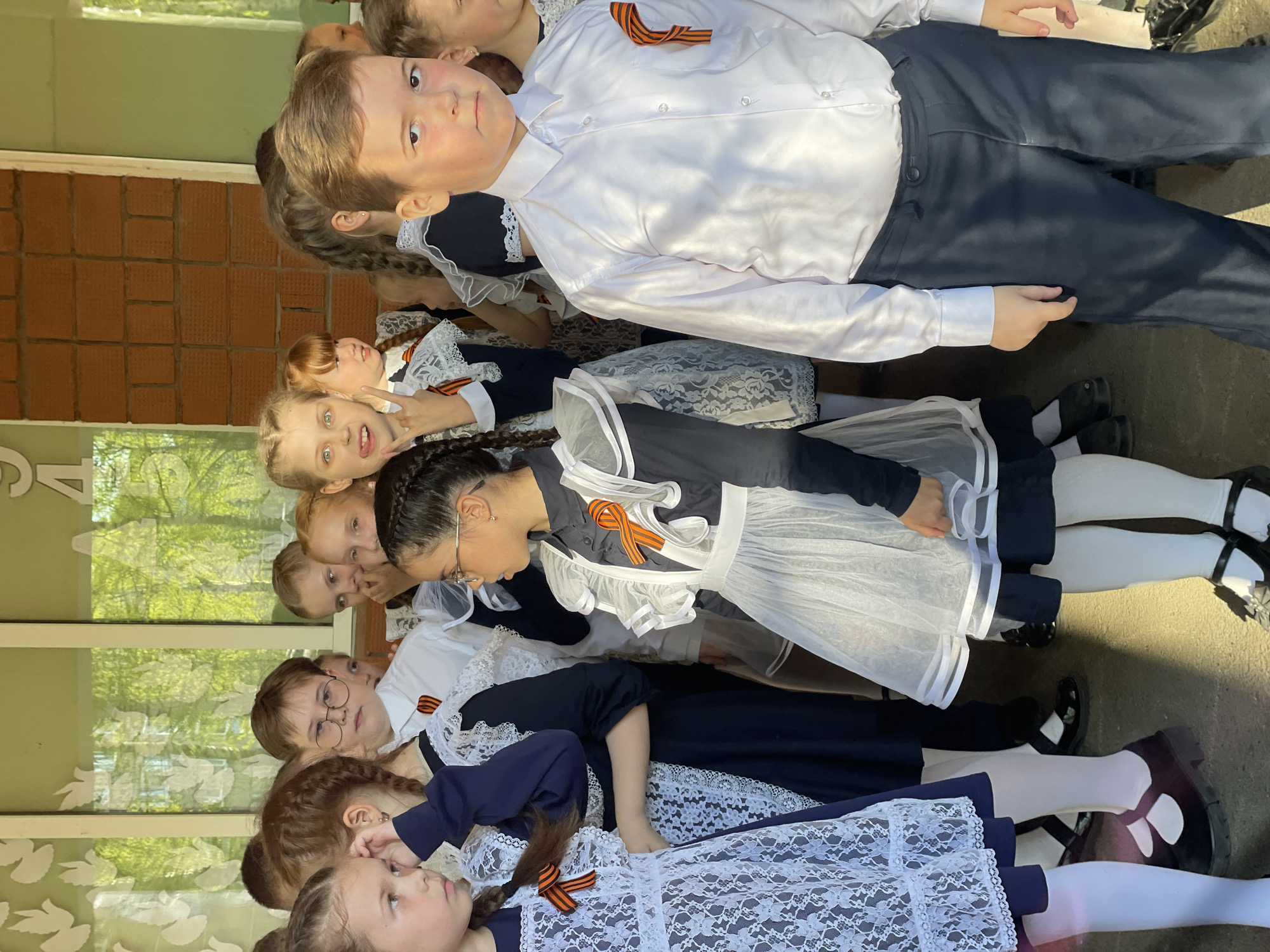 A group of children are assembled in a line outside what appears to be a school or public building. The location is suggested by the presence of bricks, windows, and a partial sign. The children consist of both boys and girls. The girls are wearing dark blue dresses with white lace aprons and white tights and black Mary Jane shoes. Many of the girls have braids. The boy at the top of the image is wearing a white shirt and dark pants. Many of the children are wearing orange and black striped ribbons, possibly St. George's ribbons. The children appear to be posing for a photo, with some looking directly at the camera.