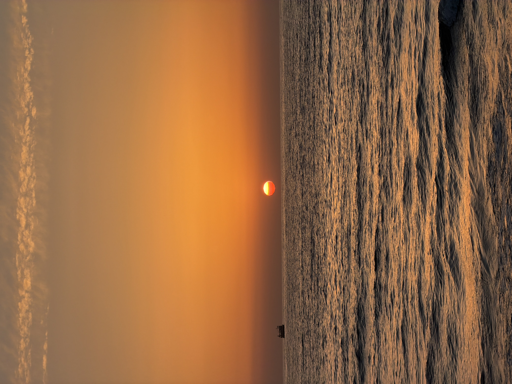 An expansive landscape image captures a vibrant sunset over the sea in Tel-Aviv, Israel. The scene is dominated by a rich palette of warm colors, transitioning from bright oranges and golds around the setting sun to softer amber and light brown hues in the sky.
The sun, a distinct orange-red orb, is positioned low on the horizon, slightly to the left of the frame's center. Its warm glow radiates across the sky, creating a smooth, atmospheric gradient that extends from the horizon upwards. A clear, horizontal band marks the horizon line, separating the radiant sky from the vast expanse of the ocean below.
The sea, filling the lower portion of the image, reflects the warm light of the setting sun with subtle golden undertones. The water's surface is highly textured, displaying countless ripples and small waves that create a detailed, almost furrowed pattern, particularly noticeable in the foreground.
In the distance, a small, dark silhouette of what appears to be a boat or a marine structure is visible directly on the horizon line, positioned beneath the sun. The upper-right portion of the sky features a few wispy, elongated cloud formations, catching the lingering light of the sunset. The overall atmosphere conveyed is one of tranquility and dramatic natural beauty. There is no visible text in the image.