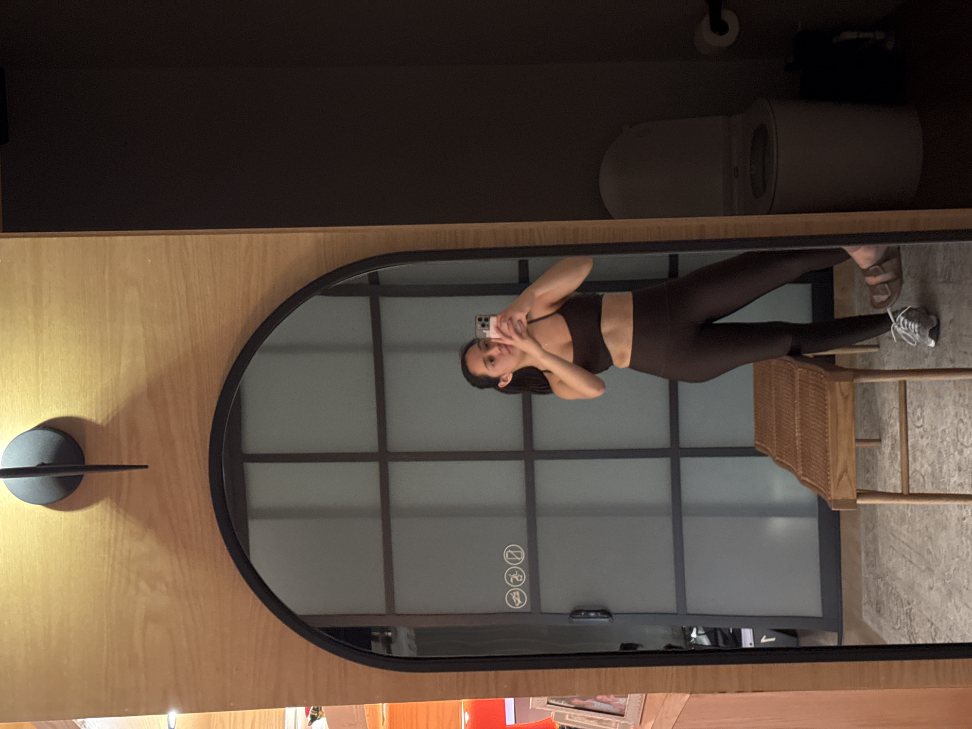 A person is taking a selfie in front of a mirror. The reflection in the mirror shows the person is wearing a dark sports bra and leggings. Behind the person in the reflection is a large window with a grid pattern, and a chair is visible to the right. The setting appears to be a room with a minimalist design. Above the mirror is a toilet. The person seems to be in a bathroom. Based on the details visible in the image, it is not possible to confirm if the image was taken in Puebla, Mexico, or to deduce the specific location.