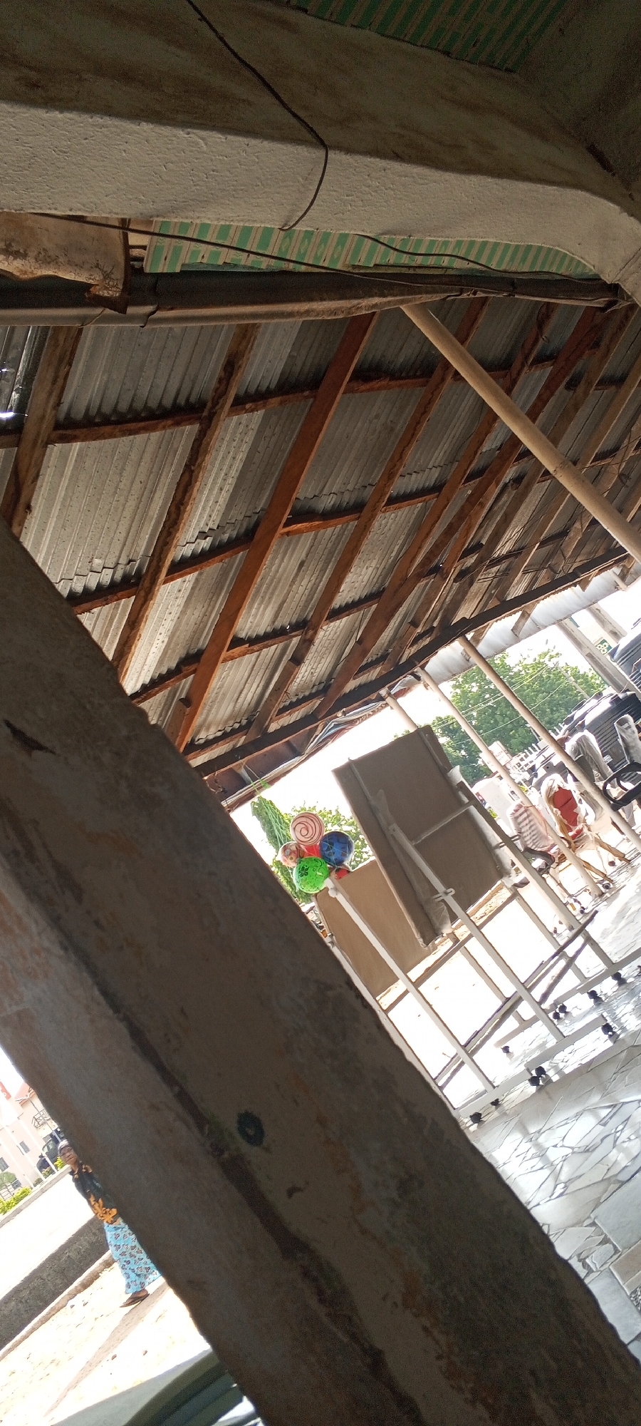 The image depicts an outdoor scene under a corrugated metal roof supported by wooden beams. In the foreground, a large, weathered wooden beam runs diagonally across the frame. In the background, through an opening, a paved area with a geometric tile pattern is visible. Several items are present on this paved area, including white easels, chairs with striped cushions, and a cluster of colorful, spherical objects that resemble balloons or large marbles. A person wearing a patterned headscarf and a blue and white skirt is standing to the left, facing away from the camera. The general environment suggests an open-air market or event space. The presence of a person dressed in a headscarf and skirt, along with the outdoor setting, is consistent with locations found in Jalingo, Nigeria.