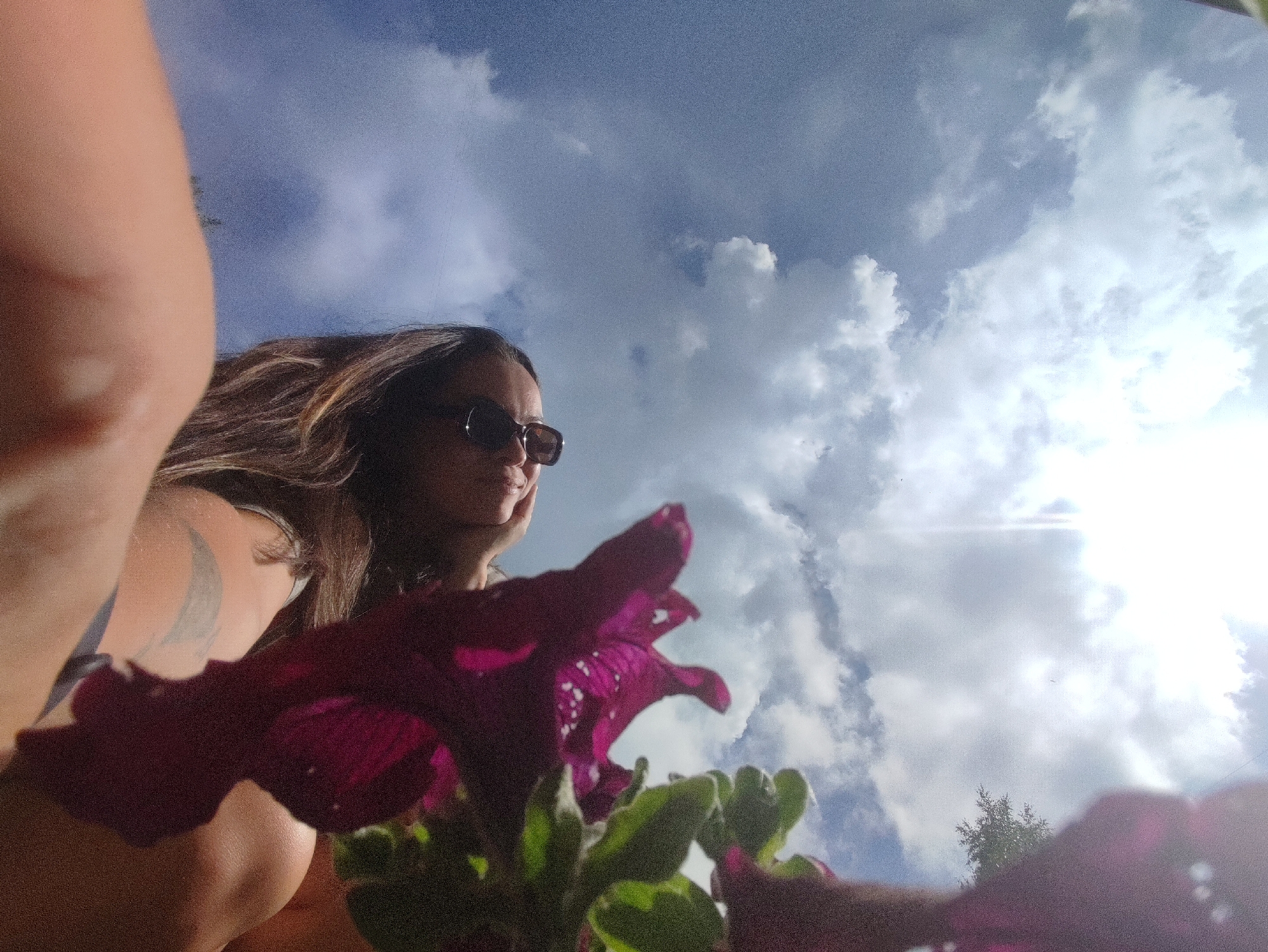 The image shows a low-angle shot of a woman with long, brown hair wearing sunglasses, looking upwards towards a cloudy sky. A large, dark pink flower is in the foreground, partially obscuring the woman's face and chest. A portion of the woman's arm and shoulder is visible on the left, with a tattoo on her upper arm. The background is dominated by a bright, cloudy sky. There are no discernible elements that place this image specifically in the city of Chaikovsky, Russia.