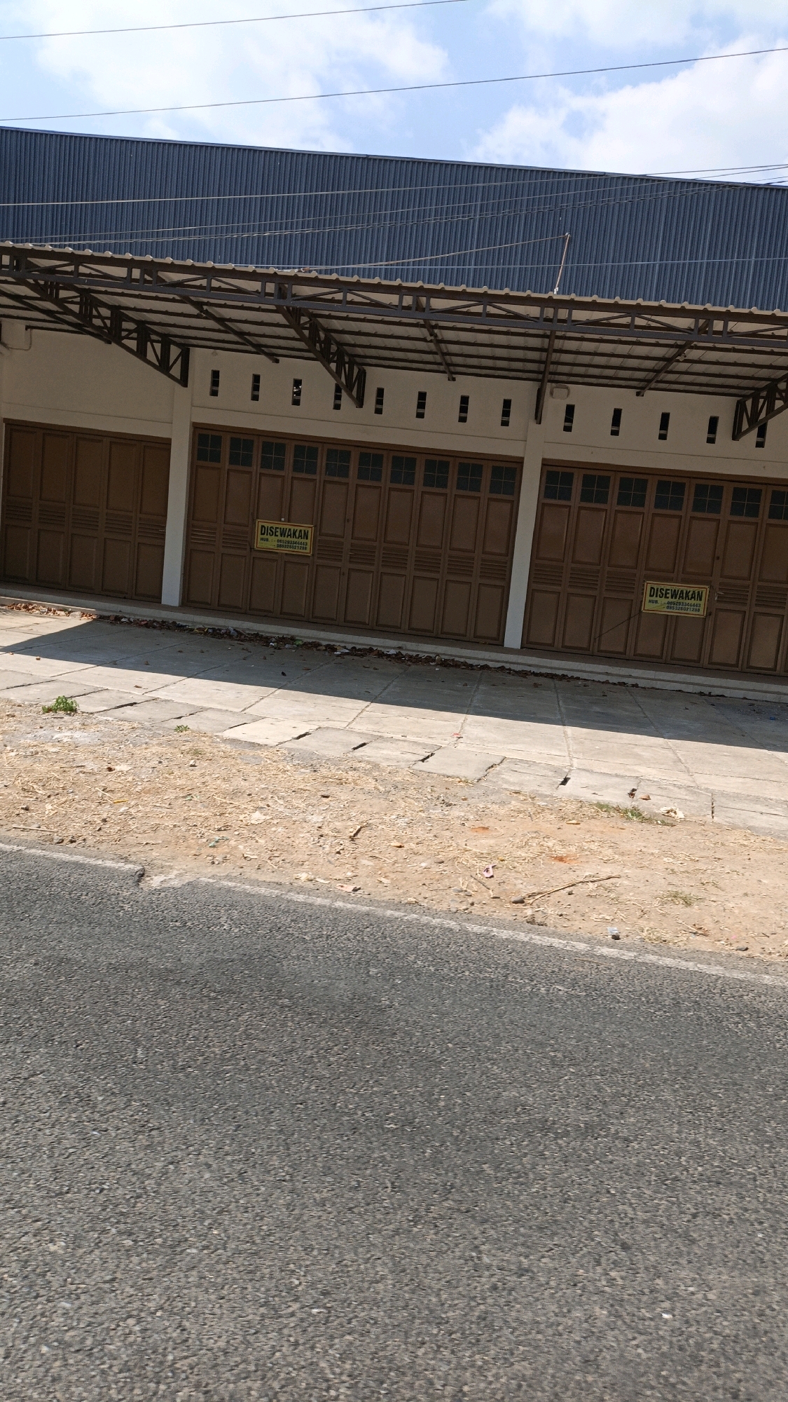 The image shows a street view in Cikal, Indonesia, featuring a row of closed storefronts with brown accordion doors. Two yellow signs, one on the left and one on the right, are visible on the doors, both reading "DISEWAKAN" (For Rent) and displaying a phone number. The storefronts have a light-colored facade and are covered by a metal awning supported by metal beams. Above the awning, a dark blue corrugated metal roof is visible. The foreground consists of a paved road, with a strip of dirt and cracked concrete sidewalk bordering the storefronts. Debris, including leaves and small branches, is scattered on the ground. The sky above is partly cloudy.