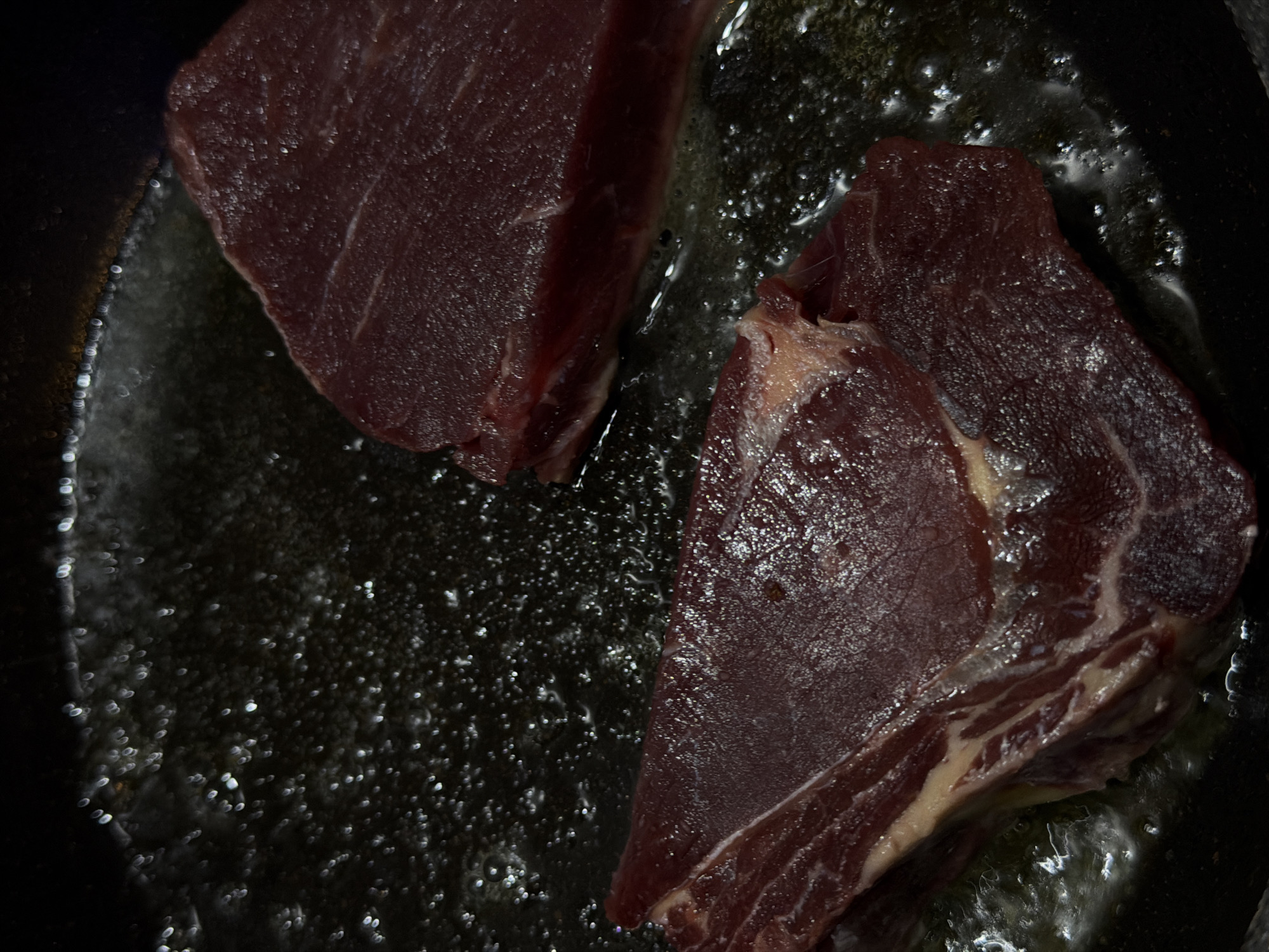 Two pieces of raw beef are cooking in a pan with oil. The oil is bubbling around the meat. The image does not contain information to confirm the location as North Kuta, Indonesia.