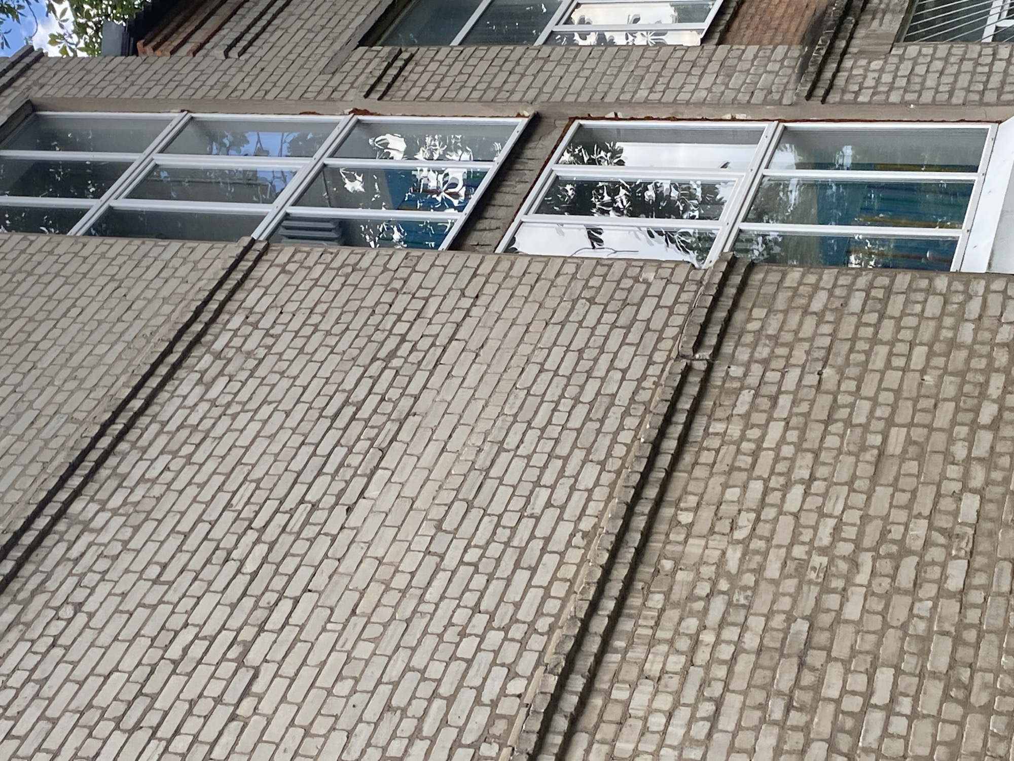 The image shows a close-up, oblique view of a weathered, light-colored brick building with several tall, multi-pane windows. The bricks appear to be laid in a stretcher bond pattern and show signs of age and dirt. A dark horizontal line, possibly a conduit or a decorative band, runs across the brickwork in the upper left portion of the image. The windows have white frames and reflect the surrounding environment, including trees and sky. Some of the windows have visible blinds or curtains inside, appearing in shades of blue and white. The architectural style suggests an apartment building or a similar urban structure. The setting is likely a residential area in Kharkiv, Ukraine, based on the provided context. No people are visible in the image.