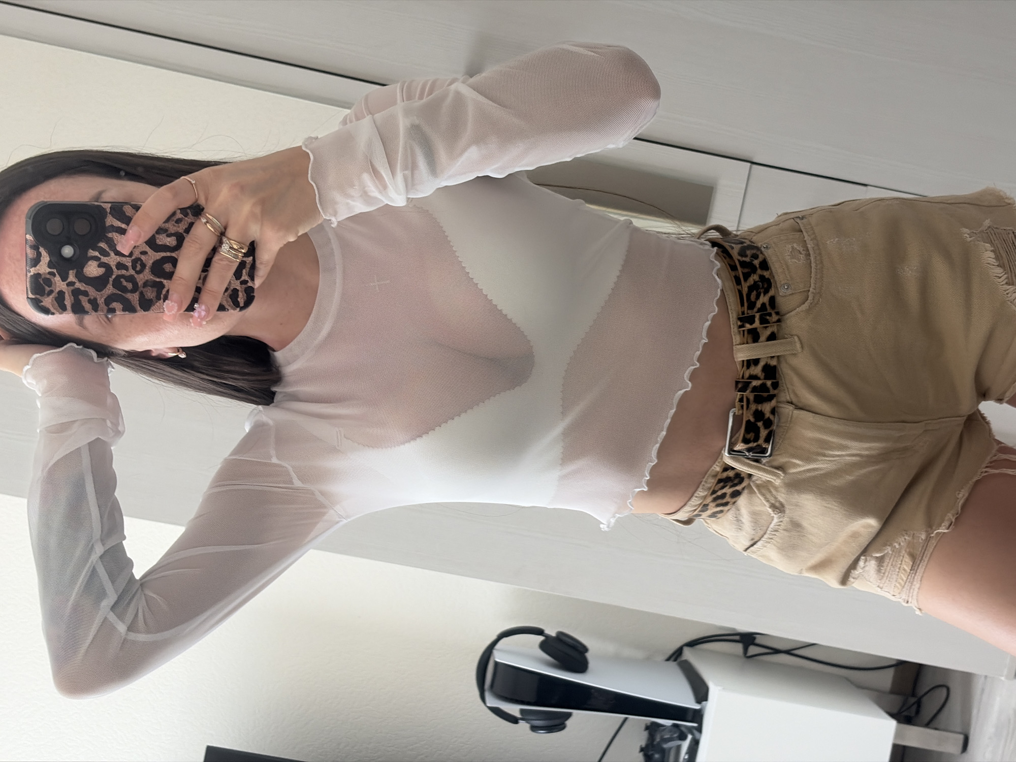 A young woman is taking a mirror selfie in what appears to be a bedroom. She is wearing a sheer white long-sleeved top with ruffled edges, a white bra underneath, and distressed tan shorts with a leopard print belt. She has her hair down and is holding her phone up to take the picture, with her right hand on her head. On the left side of the image, a white PlayStation 5 console with black headphones is visible on a white stand. The background is a plain white wall and a white door.