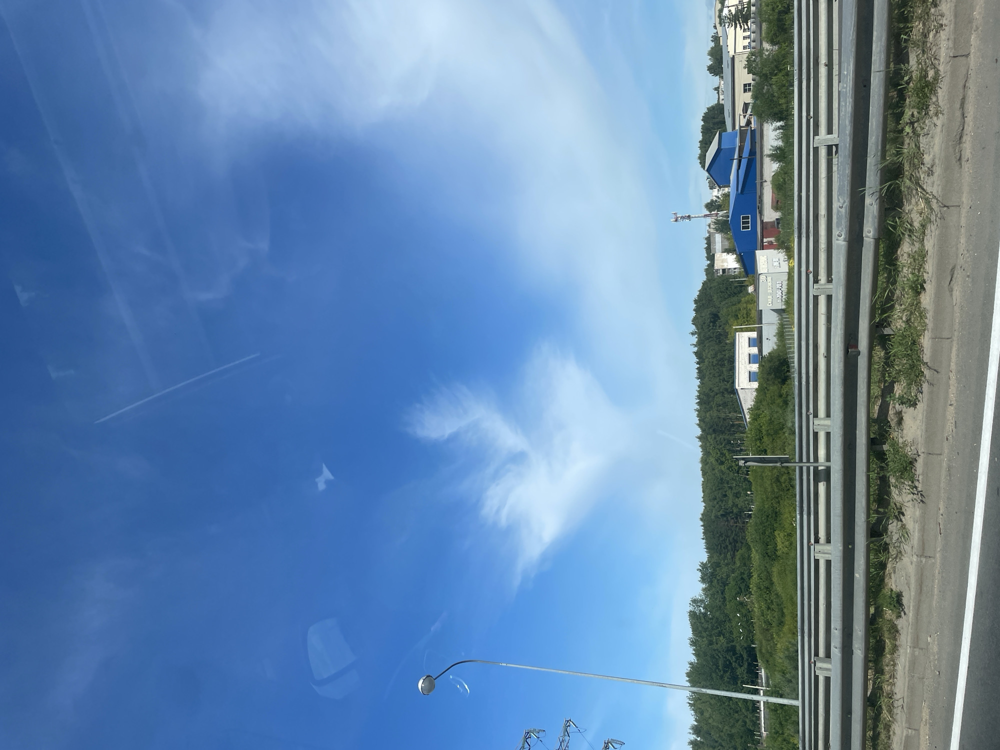 The image depicts a wide, clear blue sky with wispy white clouds and contrails. In the foreground, a metal guardrail runs horizontally across the frame, with a paved road and a strip of grass and dirt below it. A tall, silver streetlamp stands on the left side of the image. Behind the guardrail, a dense line of green trees forms the horizon. Several industrial buildings, primarily blue and white, are visible among the trees, with a telecommunications tower in the distance. The scene is viewed from a low vantage point, possibly from a vehicle on a highway. The location is identified as Rusanovo, Russia.
