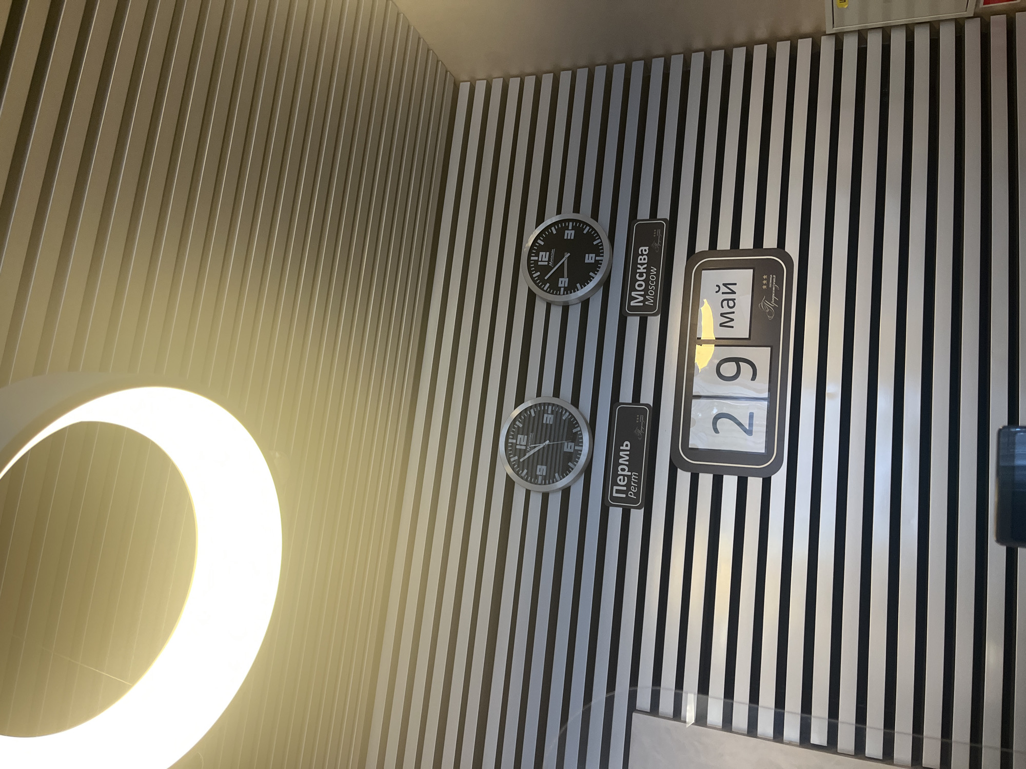 This image depicts a wall with alternating black and white vertical stripes. On the wall are two clocks, one labeled "Москва Moscow" and another labeled "Пермь Perm", along with a date display showing "9 Май" (May 9). Based on the labels, this scene is potentially located in a building or space related to Moscow and Perm.