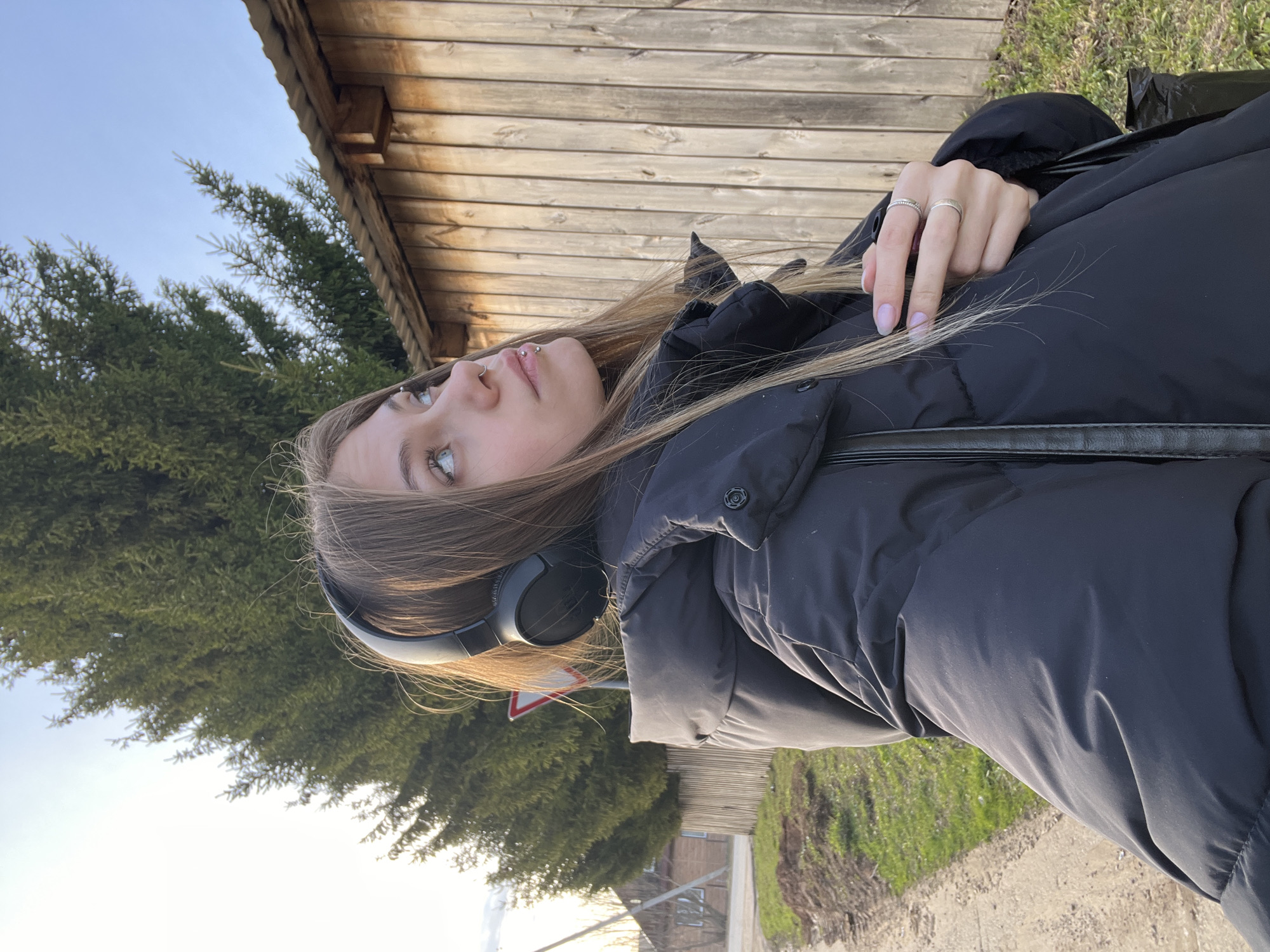 A young woman is photographed outdoors. She is wearing headphones and a dark-colored puffer jacket. She has light brown hair and fair skin. Her left hand rests on her chest, revealing two rings on her fingers. The background consists of a wooden building, trees, and a road. The photograph seems to have been taken in a rural area in Фоки, Russia.