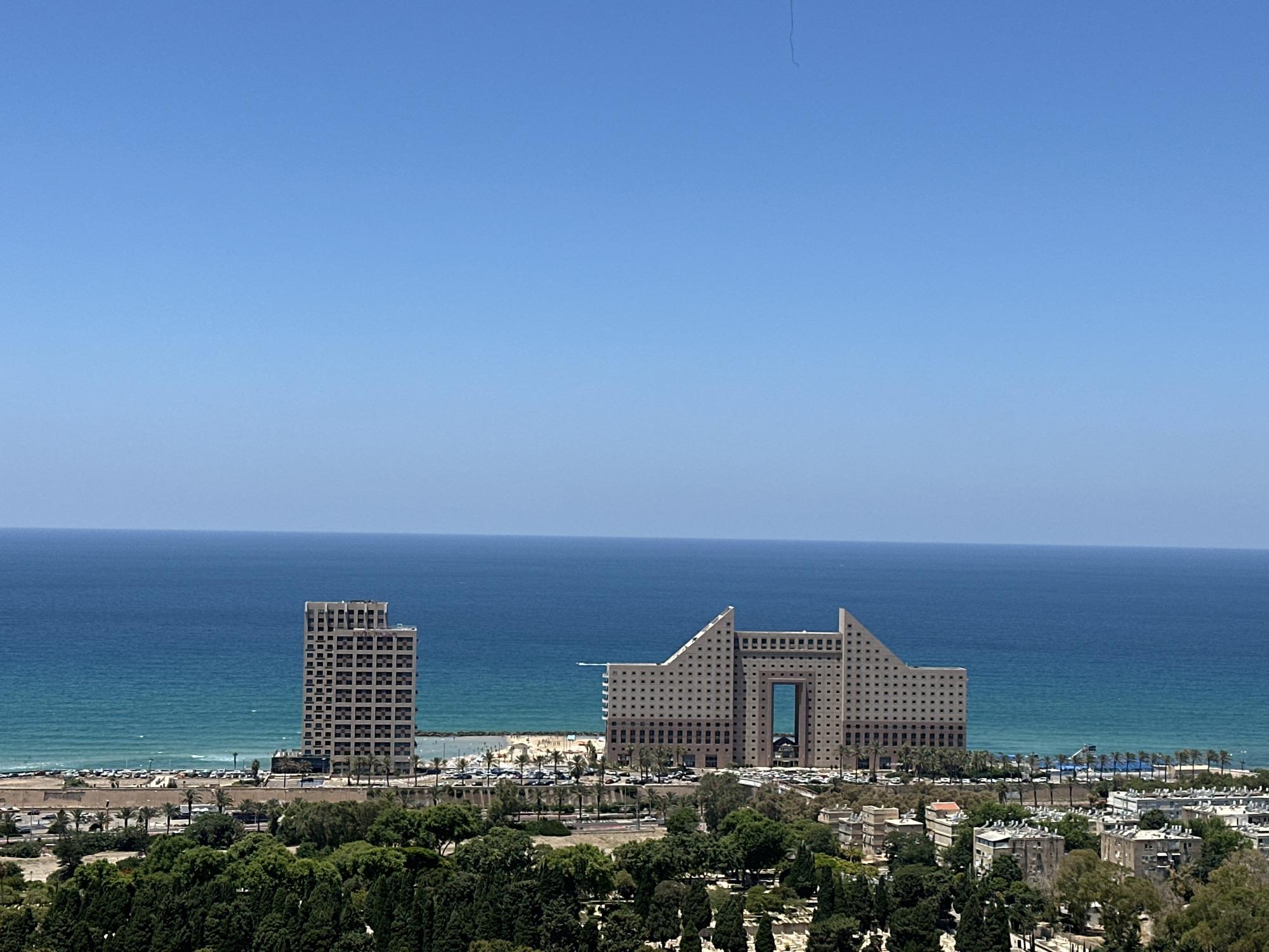 This is a wide shot of the coastline in Haifa, Israel, on a clear, sunny day. The image is dominated by the bright blue sky and the turquoise waters of the Mediterranean Sea. In the mid-ground, two distinct buildings stand out against the sea. To the left, a tall, rectangular building with a grid of windows rises from the coast. To its right, a larger, more architecturally unique building features a distinctive, angular design with a large, open archway in its center, also covered in a grid of small windows. Between these buildings and the sea is a strip of beach, some palm trees, and a parking lot filled with cars. In the foreground, a dense collection of green trees forms a natural buffer, with some buildings of a lower rise, characteristic of a city, interspersed among the foliage. The overall impression is one of a coastal city with modern architecture set against a backdrop of natural beauty.