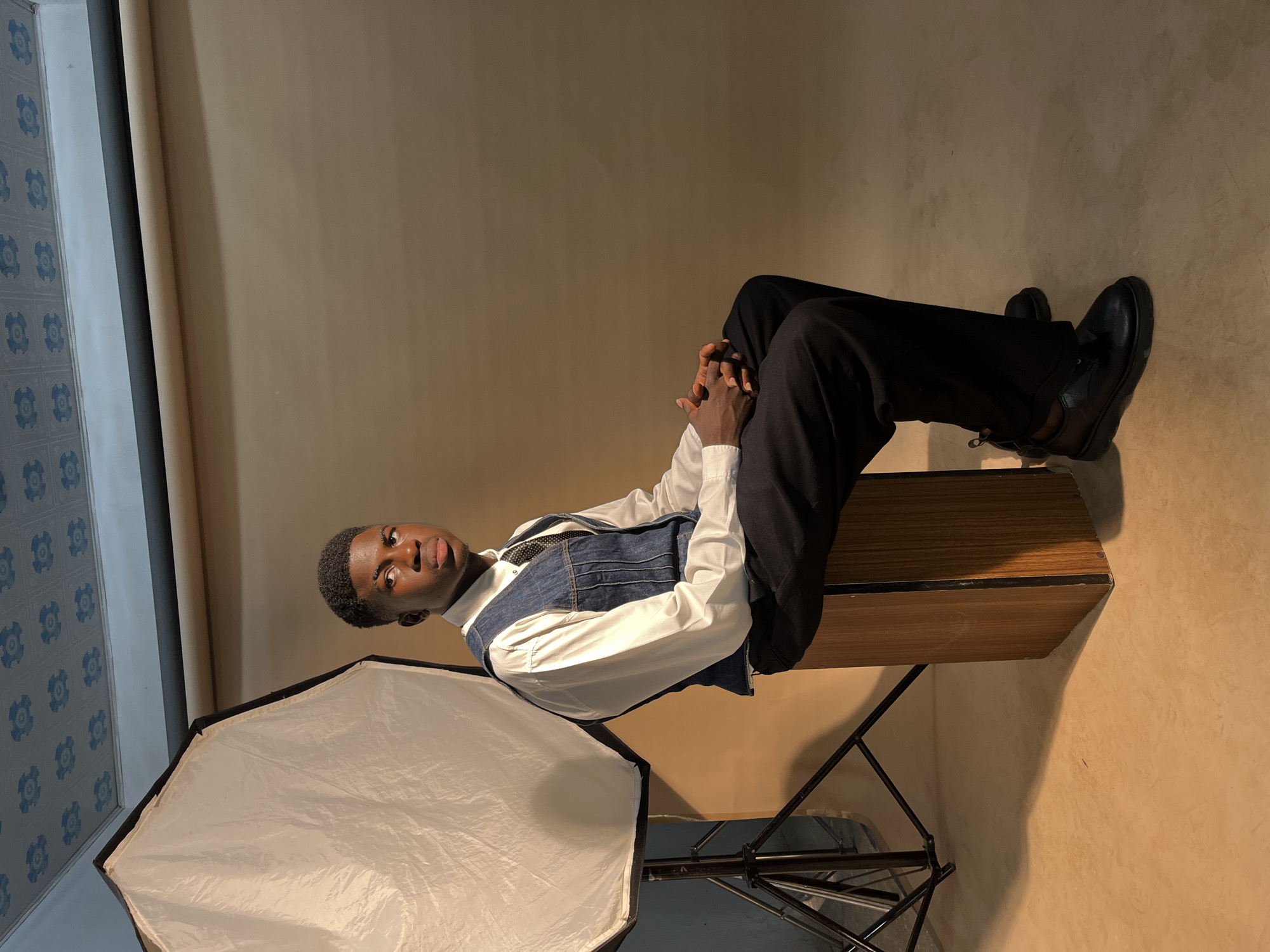 The image shows a young Black man seated in a studio setting, likely in the city of Unknown, Unknown. He is posed on a wooden structure with black metal legs, possibly a bench or small table, and is facing the camera at an angle. He wears black trousers, black leather shoes, a white collared shirt, and a denim vest. His hands are clasped together. A softbox is positioned to the lower left of the frame, providing lighting. The backdrop consists of a plain, light beige wall and a section with a blue and white patterned wallpaper or decorative panel. The overall lighting in the studio appears to be a combination of natural and artificial sources.