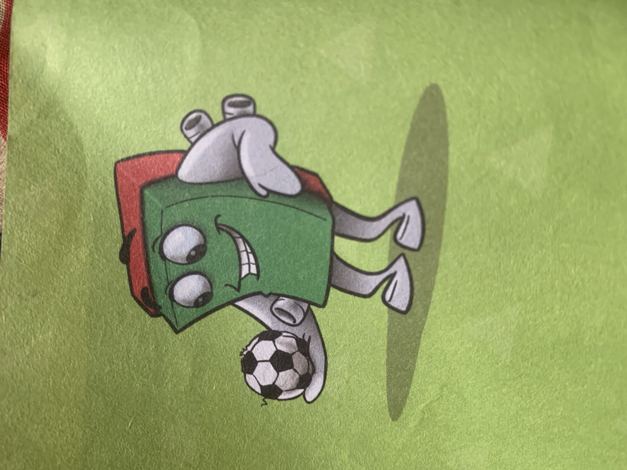 The image shows a cartoon drawing of a green anthropomorphic book wearing a red cap and a backpack. The book character is standing on a green surface, which appears to be a field, and is holding a soccer ball in its left hand. The character has large, cartoonish eyes, a wide smile, and two white arms and legs. The background is a solid, textured green. There is a shadow cast on the ground behind the character. The image does not contain any information about the city of Jauro Yinu, Nigeria.