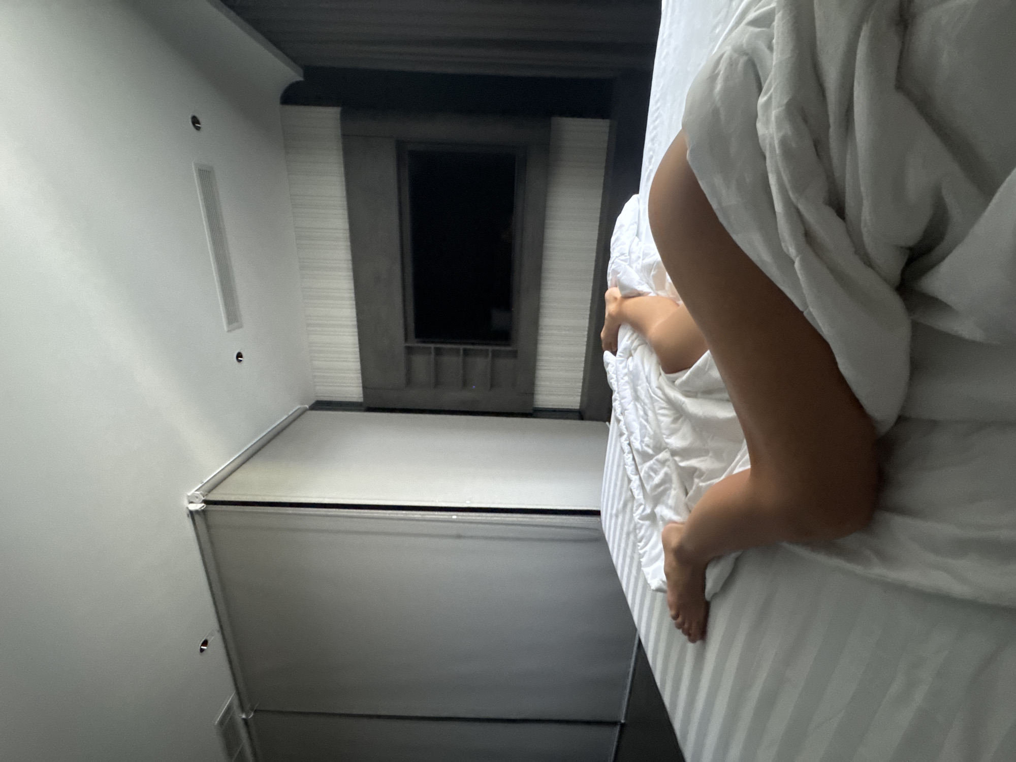 A low-angle shot inside a room shows a bed with white sheets. On the bed, a person is lying under a white duvet. Part of their bare leg and foot are visible. The room has a white wall with a vent, and what appears to be a closed cabinet or drawer system. A dark, rectangular panel (possibly a TV screen) is framed by textured, vertical panels. The lighting suggests it is daytime indoors. The location is unspecified.