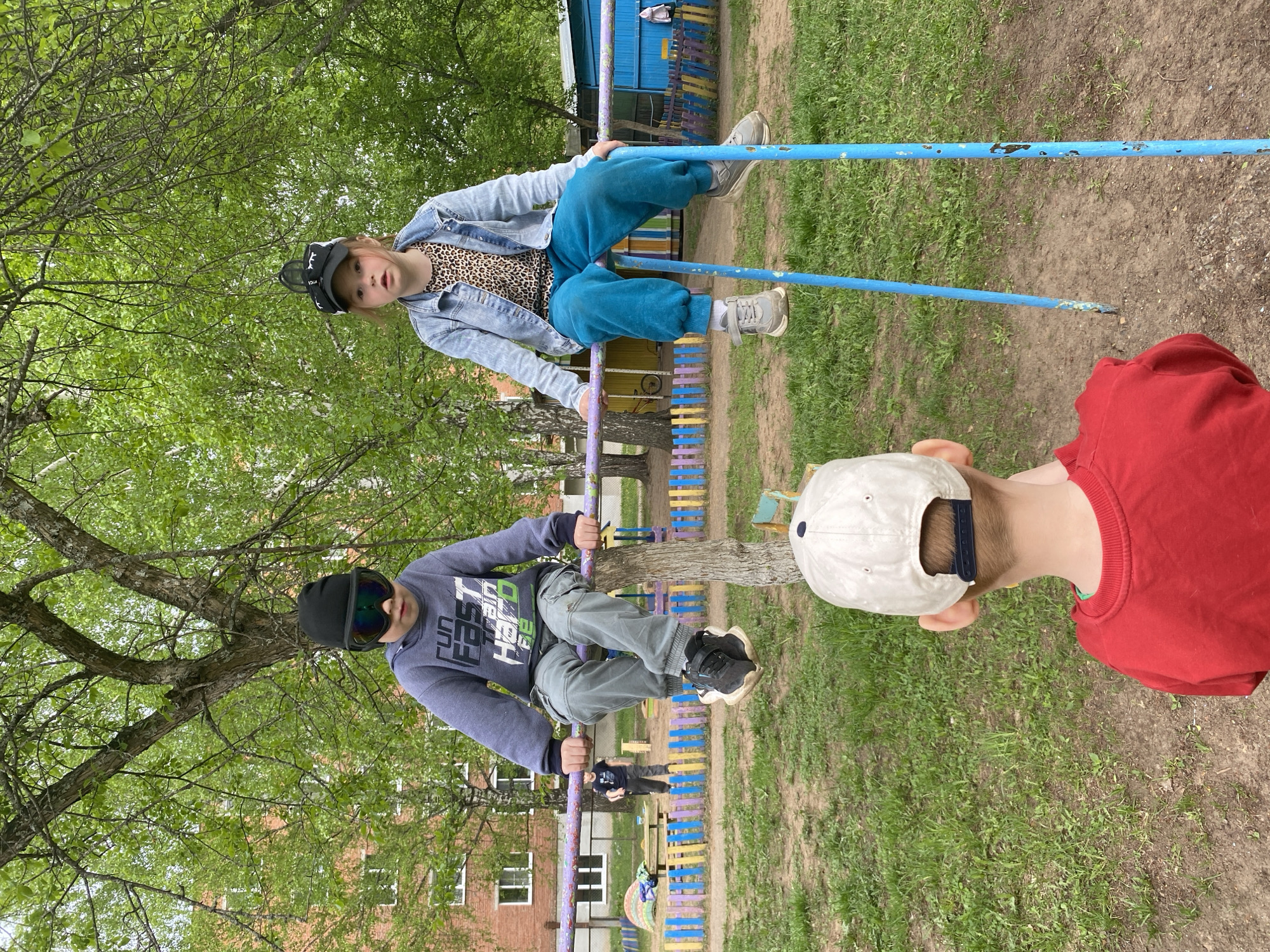 Two children are positioned on parallel blue bars in an outdoor play area in Noviy, Russia. The first child wears a denim jacket over a leopard print shirt, teal sweatpants, a black baseball cap and grey sneakers. The second child wears a grey sweatshirt with graphic text, grey sweatpants, black sneakers, a black hat and sunglasses. A third child, wearing a red shirt and white baseball cap, faces away from the camera. The play area includes a colorful fence and building visible in the background. Trees surround the play area.