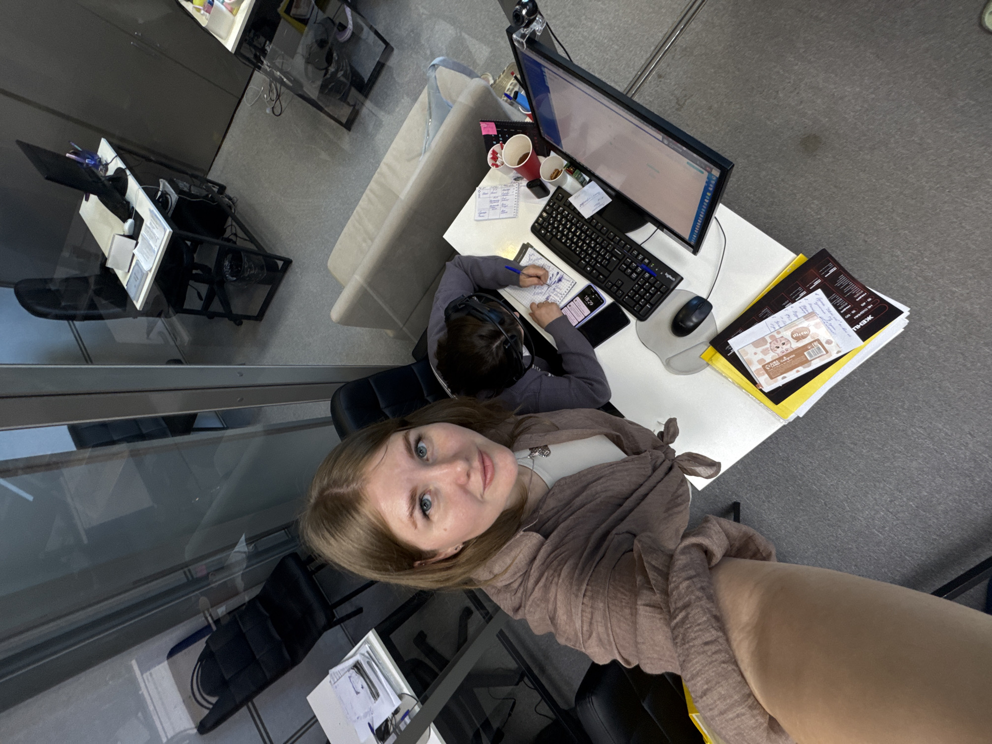 A woman is taking a selfie in an office. Another person, wearing a headset, is seated at a desk with a computer, keyboard, mouse, and papers. The desk also has cups and a notebook. The location is potentially in Perm, Russia, based on the context of the user's query.