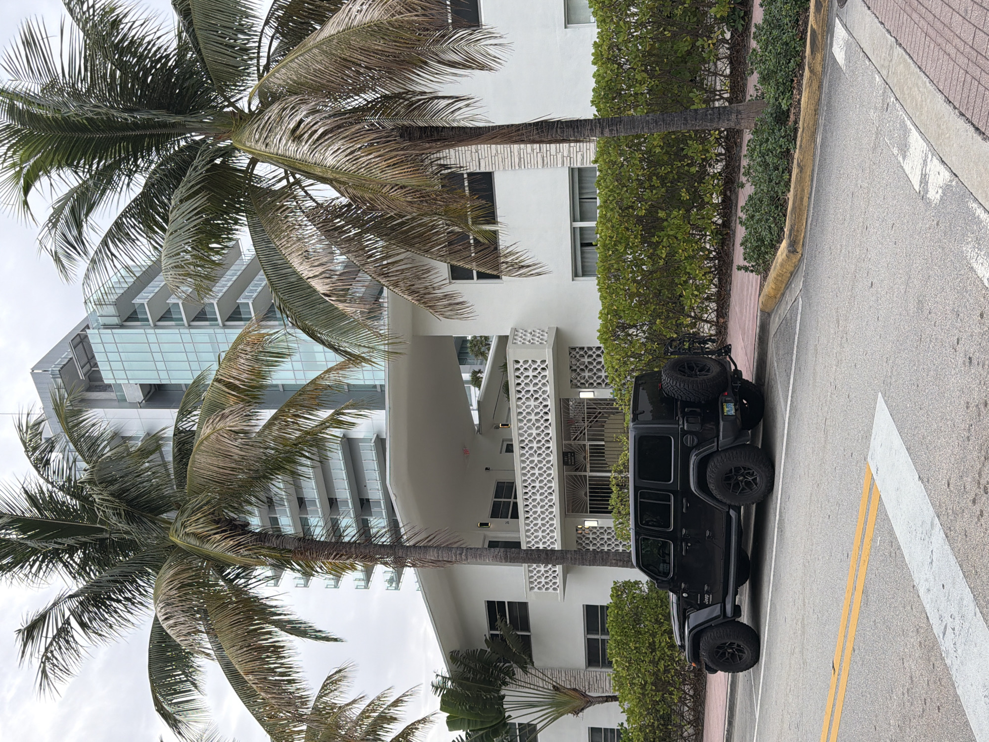 A black Jeep Wrangler is parked on the street in front of a white Art Deco building in Miami Beach, Florida, USA. The building has a white, curved overhang and a white decorative railing on the second floor. Palm trees are visible in front of the building and on the right side of the image. A taller, modern glass building is visible behind the Art Deco building. The street has yellow and white lines painted on it, and a pedestrian walkway made of red brick is visible to the right of the parked Jeep. The sky is overcast.