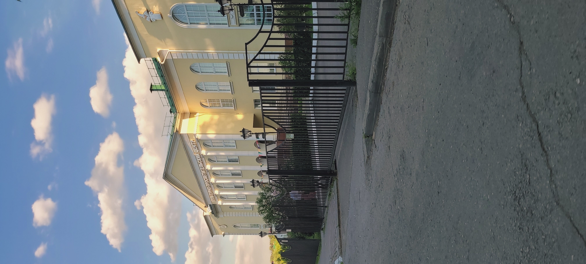 The image displays a large, pale yellow classical or neoclassical style building with a green roof, situated under a partly cloudy blue sky in an unknown city and region. The building features a prominent central pediment supported by several white columns, with the text "КУЛЬТУРНО-ПРОСВЕТИТЕЛЬСКИЙ ЦЕНТР" (Cultural and Educational Center) inscribed on its facade below the roofline. Numerous white-framed windows, primarily tall and arched, are visible across the building's front. Decorative architectural elements, resembling volutes, are present above some windows on the right side. Two ornate black lampposts are positioned in front of the building, behind a security fence.
In the foreground, a dark asphalt road with minor cracks occupies the lower portion of the frame. A grey concrete curb separates the road from a paved area directly in front of a tall, black wrought-iron fence. This fence, composed of vertical bars, extends across the width of the image and appears to be a large gate system, partially obscuring the view of the building's immediate surroundings.
Behind the fence, greenery including bushes and trees is visible. One person is partially visible walking left-to-right behind the fence, to the left of the center, wearing a vertically striped shirt (possibly blue and white or red and white) and dark pants. The scene is illuminated by strong, warm sunlight, indicating either late afternoon or early morning, which casts shadows and highlights the building's facade and the white cumulus clouds scattered across the sky.