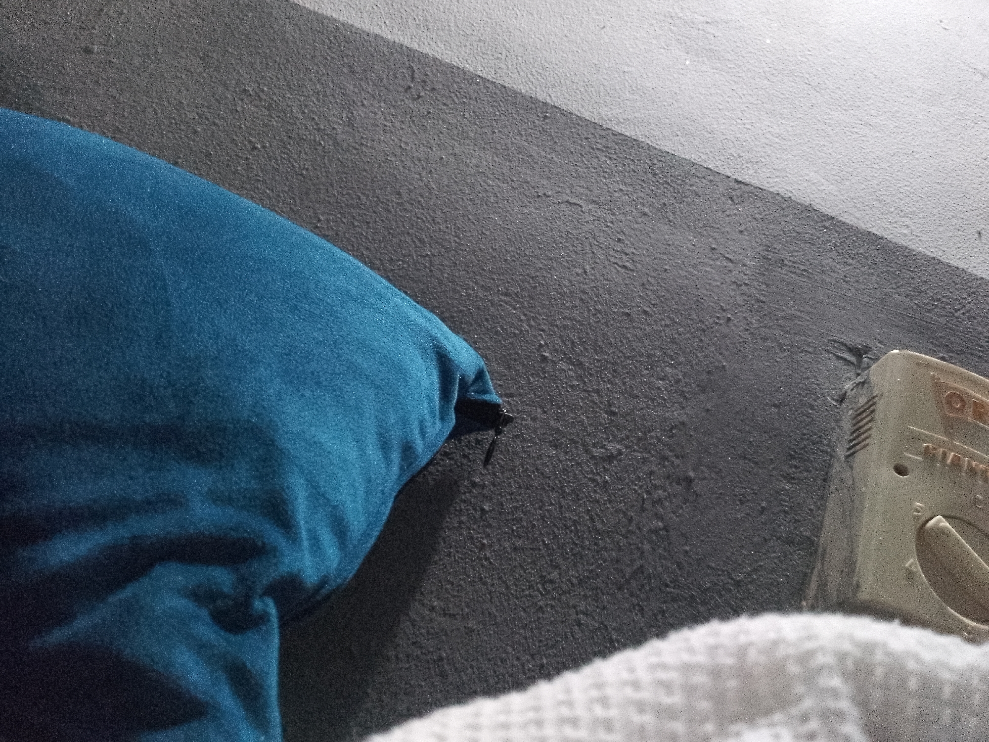 This image depicts a close-up view of a textured surface, likely a wall, with a diagonal division of color. On the left side, a portion of a teal-colored pillow is visible, its fabric appearing soft and slightly wrinkled. To the right of the pillow, the textured surface transitions from a dark gray to a lighter gray, and then to a white area in the upper right quadrant. A small, off-white electrical outlet or cover is present on the right side of the image, featuring some visible markings and a dial. In the bottom right corner, a section of a white, textured fabric, possibly a blanket or towel, is visible. The lighting suggests an indoor environment, with shadows cast by the pillow onto the textured surface. No people or specific landmarks of Jauro Yinu, Nigeria are identifiable within this frame.