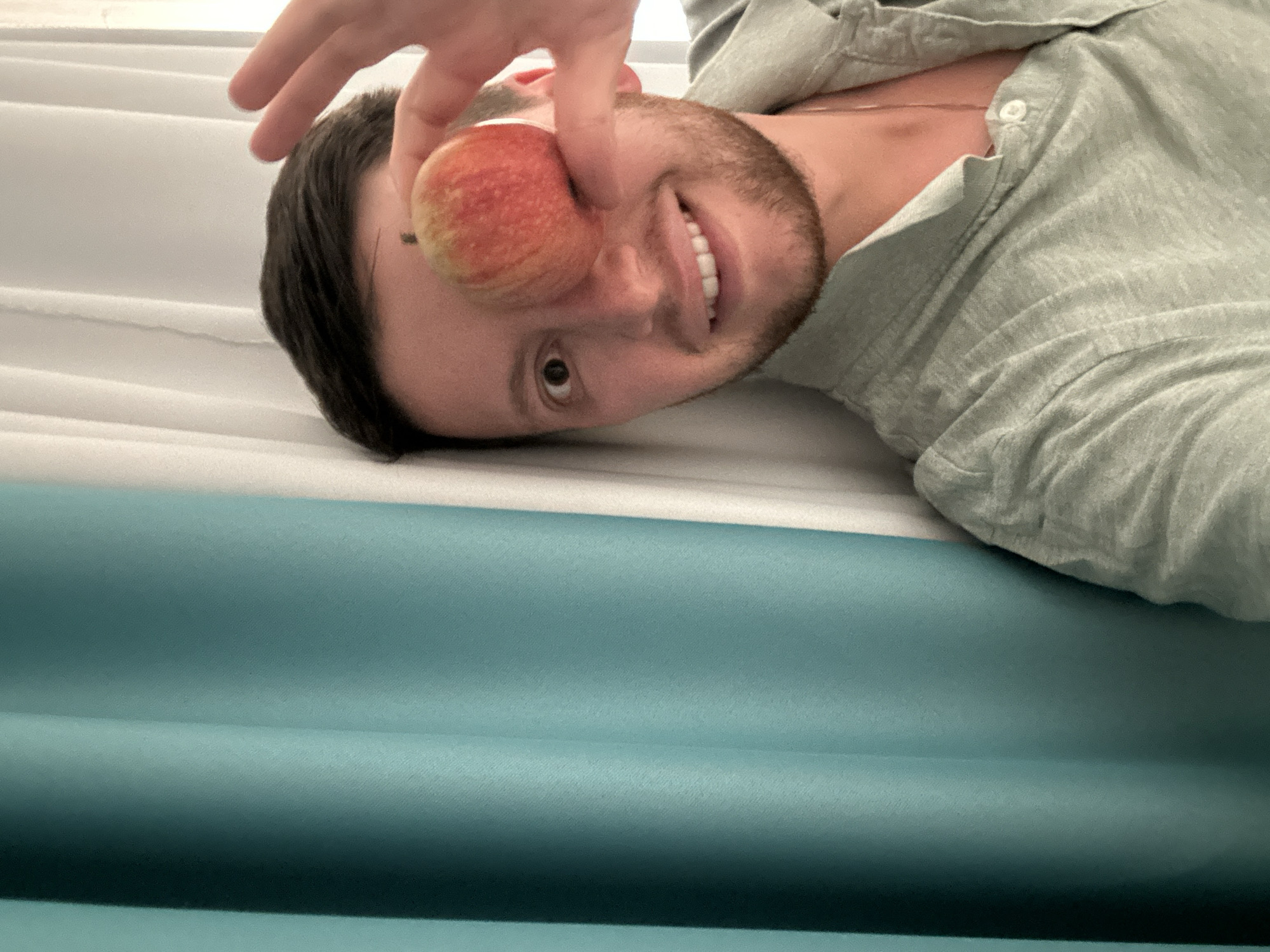 A man lies on his side on an inflatable mattress that is turquoise in the lower half of the picture and white in the upper part. He is holding a small red apple over his eye, and smiling, wearing a light-green button-up shirt. The location is unknown and there is no indication of Мирный, Russia in the picture.