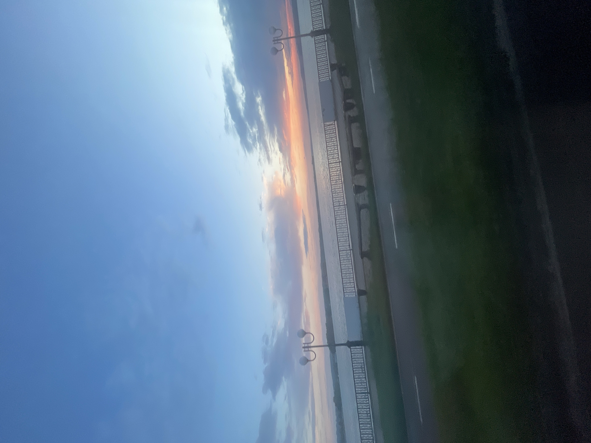 The image depicts a waterfront scene at dusk in Votkinsk, Russia, along the Kama River. The sky is a gradient of blue and soft pink and orange hues, indicating the sunset. A calm body of water, presumably the Kama River, stretches across the middle ground. Along the riverbank, there is a paved walkway with white lane markings, bordered by a railing. Two lampposts with double spherical lights are visible along the walkway, casting a soft glow. In the distance, the opposite bank of the river is lined with trees and buildings. The foreground is dominated by green grass and a dark asphalt surface, possibly a road or path. No people are present in the image. The overall atmosphere is serene and peaceful, characteristic of a quiet evening by the river.
