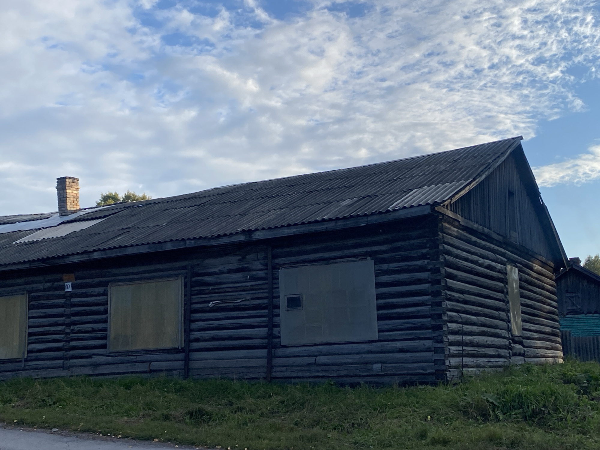 This is a full, eye-level outdoor shot of an old, dark wooden log building under a partly cloudy blue sky in Listvyanskiy, Russia. The building has a dark, corrugated roof and several boarded-up windows. A brick chimney protrudes from the left side of the roof. The building is covered in horizontal logs. A small white sign with the number "10" in blue is visible on the left side of the building. The building is situated in a grassy area next to a paved road. In the background, another building and a turquoise fence are visible.