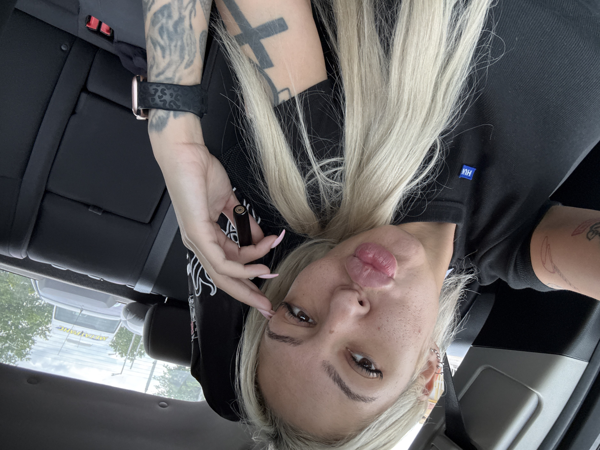 A selfie taken from within a car. The individual has blonde hair, light skin with freckles, and is wearing a black top. Visible tattoos adorn both arms. They are holding a vape pen. The car appears to be parked outside a building in Plovdiv, Bulgaria, as a reflection in the car window displays a sign with Bulgarian text ("металснаб"). The interior of the car is black, with visible car seat and seat belt components.