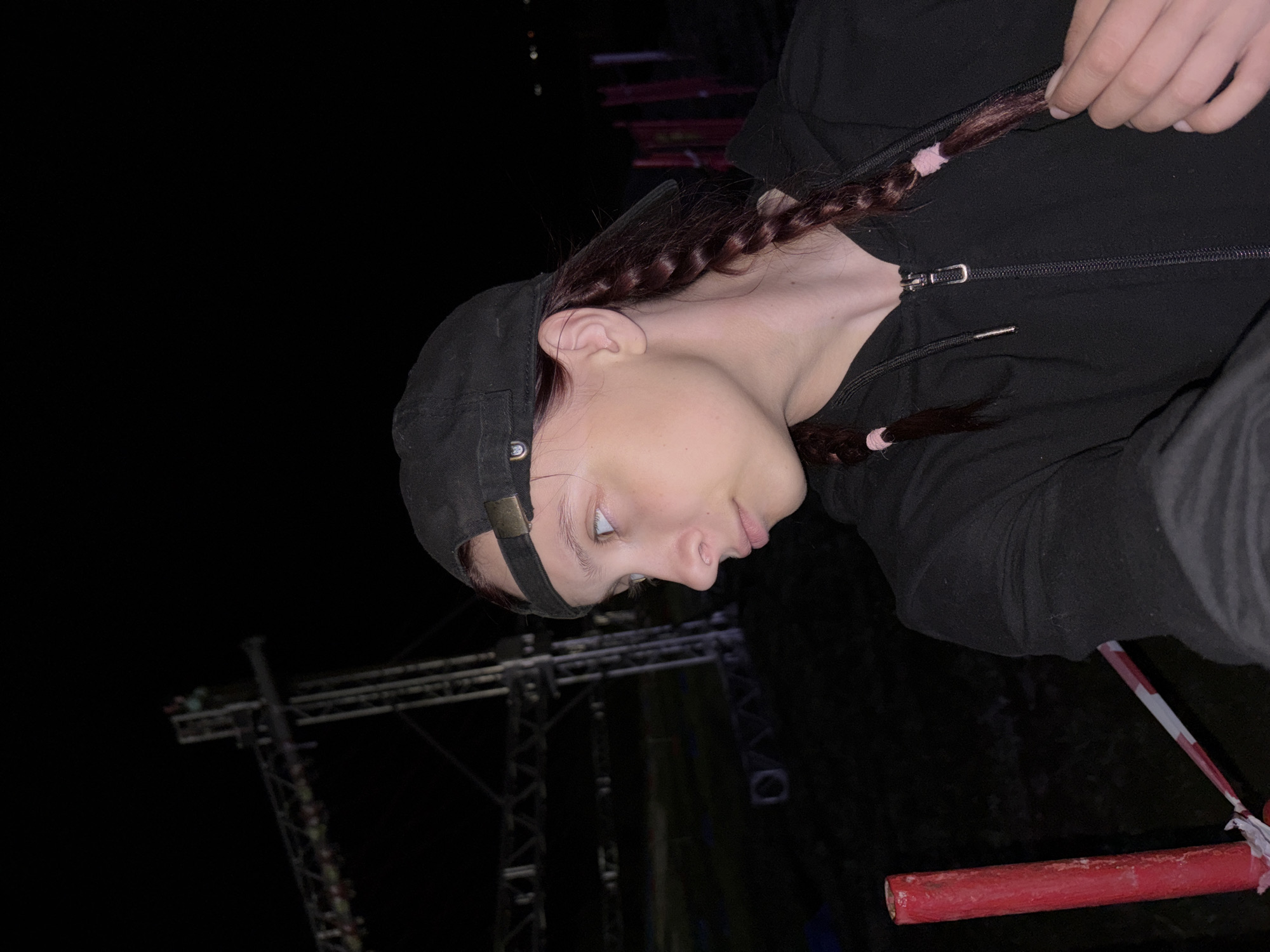 The image shows a person with light skin wearing a black hooded jacket and a dark baseball cap. The person has brown hair styled in two braids, each secured with a light-colored band. The person's face is angled slightly downward and to the right, with their eyes open. The background is dark, but a metal structure with multiple supports is visible. A red and white striped pole is in the lower right corner. The location is difficult to ascertain from the image alone, and there is no definitive evidence to place the scene in Khvalynsk, Russia.