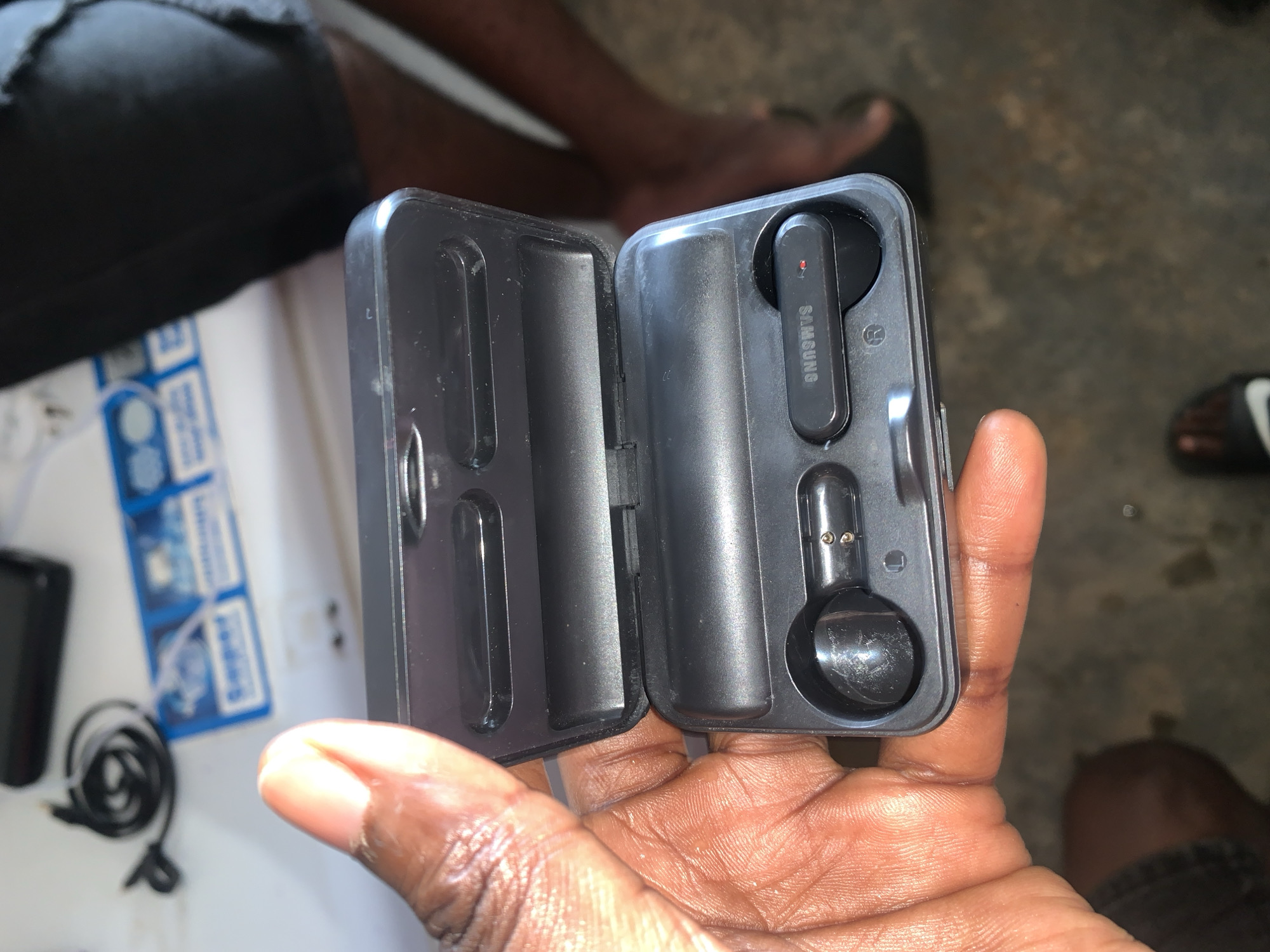 A close-up, overhead shot displays a dark-skinned hand holding an open black wireless earbud charging case. The rectangular case, with a hinged lid, reveals two black wireless earbuds resting in molded compartments. Each earbud features the "SAMSUNG" brand name printed vertically in white. The upper earbud shows a small red indicator light.
In the blurred background to the left, a white surface is visible, holding a coiled black cable (likely USB) and a black smartphone or similar device. Behind these items, blue and white packaging with partial text, including what appears to be "Supergel," can be seen. The far background shows blurred human legs and feet; one foot, wearing a dark sandal with a white swoosh logo (resembling Nike), rests on a dark, possibly concrete or tiled, ground. The natural lighting suggests a daytime setting. The image captures a moment where the earbuds are being held or presented, likely within the casual environment of the New Stadium, Nigeria.
