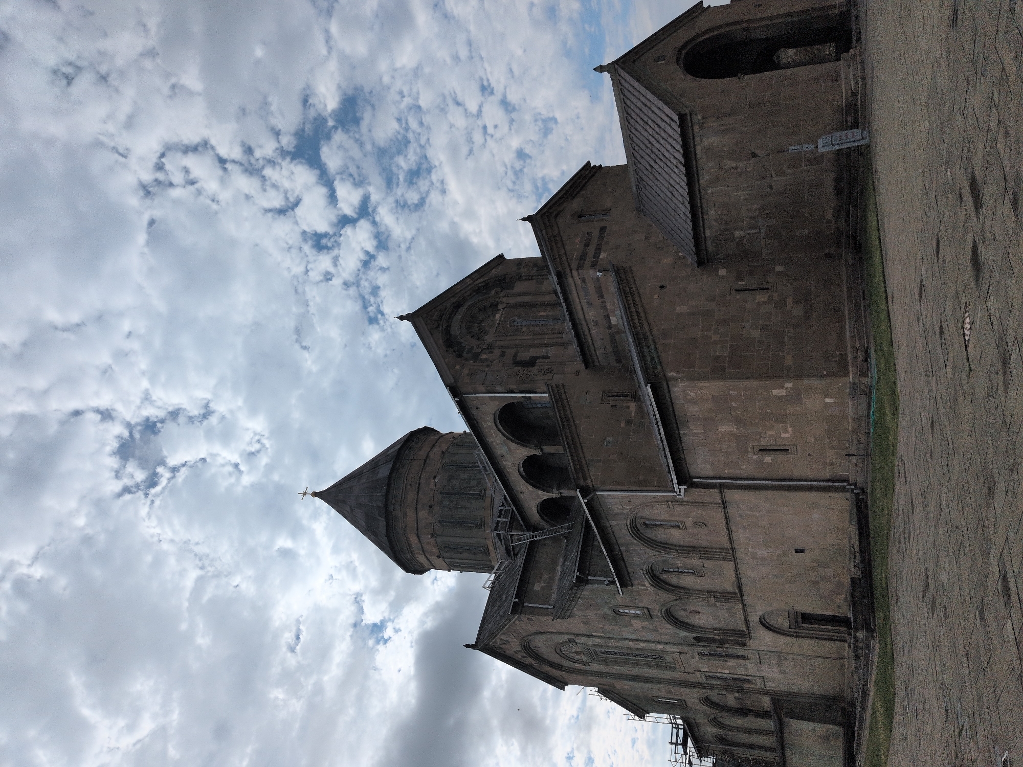 The image shows the Svetitskhoveli Cathedral, a prominent landmark in the city of Mtskheta, Georgia. The cathedral is made of stone and features a large conical dome topped with a cross. The facade of the building is adorned with arched windows and intricate carvings. The sky above is filled with dramatic clouds, casting shadows on the ancient structure. In the foreground, a paved courtyard leads to the cathedral's entrance, with a patch of green grass bordering the stone pathway. The overall scene conveys a sense of historical significance and architectural grandeur.