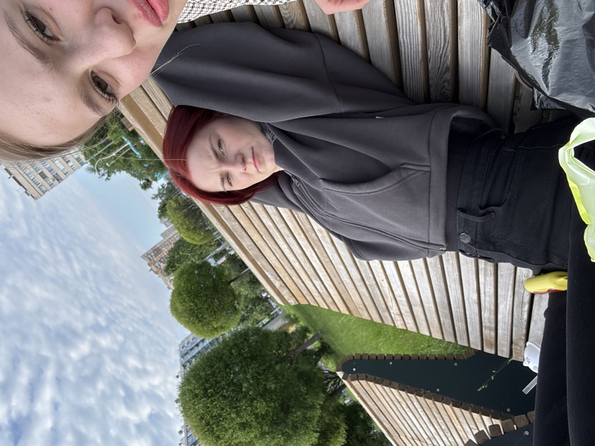 The image shows two young women sitting on a wooden park bench. The woman in the foreground, on the right, is taking a selfie, with her face close to the camera. She has blonde hair and is wearing a patterned shirt and a dark jacket. The woman in the background, on the left, has red hair and is wearing a dark gray hooded jacket and black pants. She is leaning back on the bench with her arms behind her head, looking off to the side with a neutral expression. Behind the women, the scene includes a grassy area, manicured trees, and apartment buildings in the distance, suggesting an urban park setting. The sky is partly cloudy. A black trash bag is visible on the bench to the right of the woman in red.