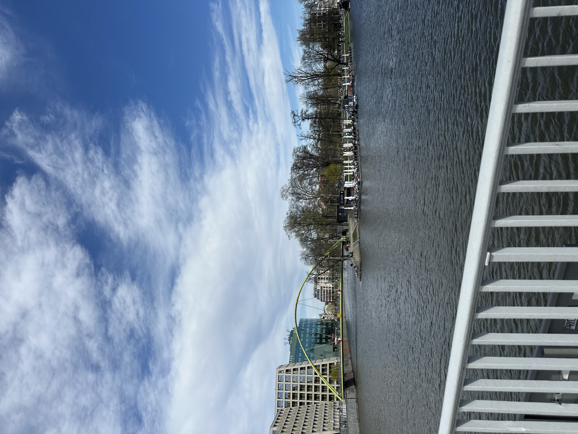 The image presents a view of Wrocław, Poland, from a bridge. The sky is blue with streaks of cirrus clouds. A river flows through the scene, flanked by trees and buildings. A notable green arched bridge spans the river. Vehicles are parked along the riverbank. Some people are visible on the pedestrian path. The bridge railing from which the photo is taken is visible in the right foreground.